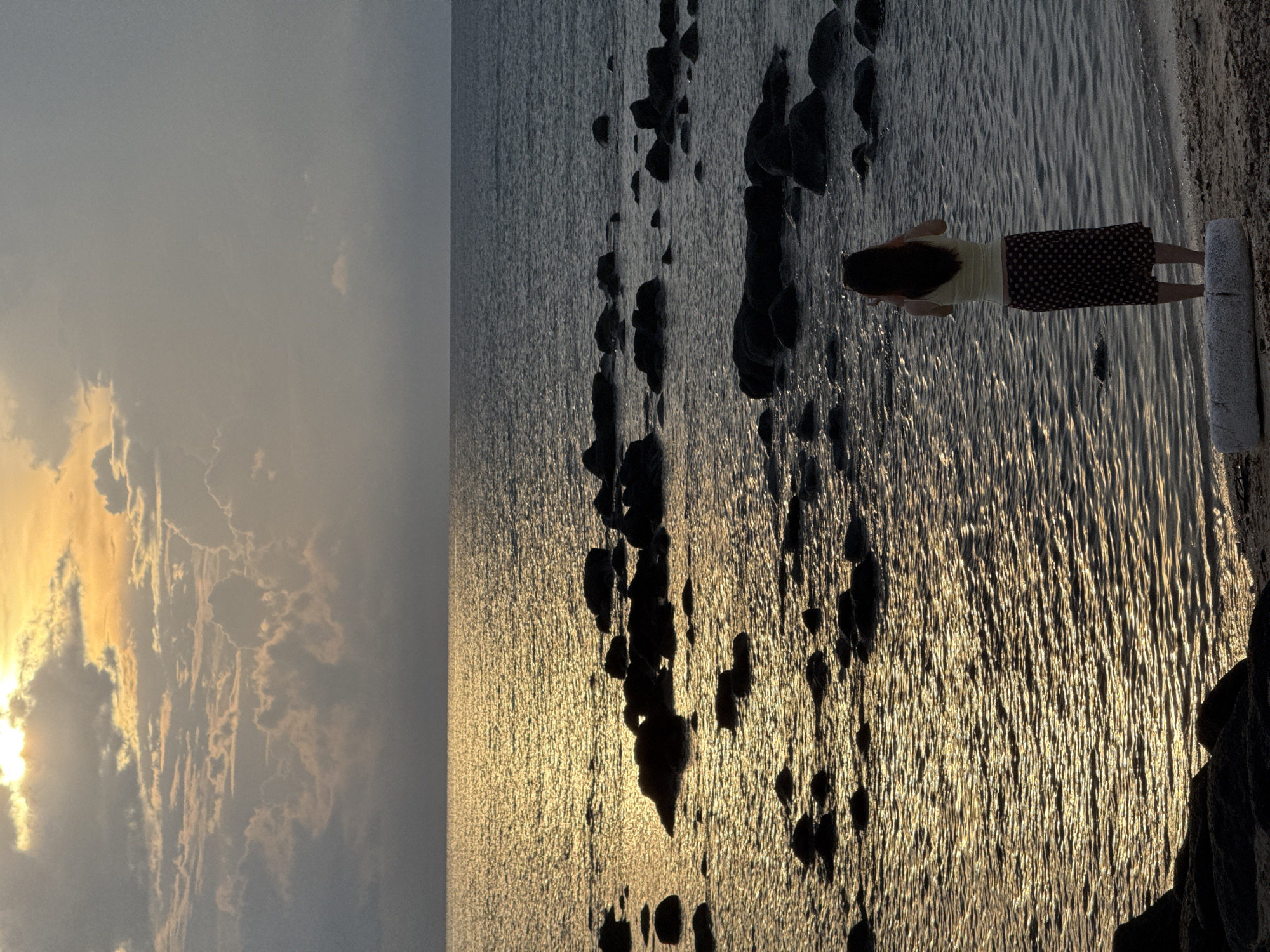 A lone woman stands on a sandy shore, facing the ocean, capturing the scene with her phone. The sun, partially obscured by clouds, casts a warm, golden glow across the water, creating a shimmering effect. Rocks are scattered in the shallow water, some partially submerged. The sky is a blend of grey clouds and patches of orange and yellow from the setting sun. The woman is wearing a light-colored top and a dark, polka-dotted skirt. She is positioned near a log on the beach. This is likely a coastal area on Lý Sơn Island, Vietnam, known for its scenic beaches and volcanic rock formations.