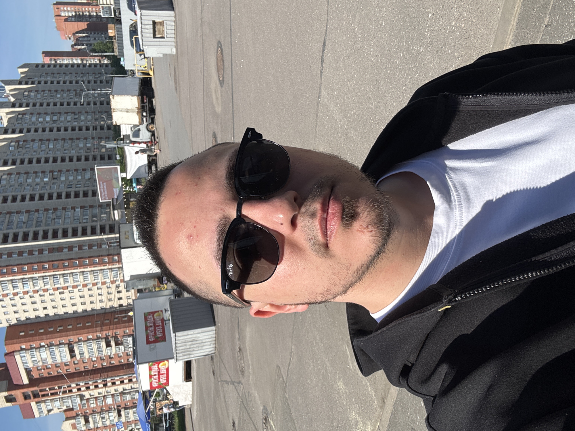 A selfie captures a man outdoors in a sunlit urban environment. The man, positioned prominently in the foreground, looks towards the camera. He wears dark sunglasses, a white crew-neck t-shirt, and a black zippered hoodie or jacket. His hair is dark and short, and he has visible stubble or a short beard. The "Ray-Ban" brand logo is faintly discernible on the frame of his right sunglass lens (on the viewer's left). Small blemishes are visible on his forehead.
The background reveals a city street or large paved area, possibly a parking lot, stretching behind him. To the left, several tall, multi-story buildings with numerous windows, featuring grey and reddish-brown architectural elements, line the horizon. In the middle ground, various vehicles, including a white box truck, are parked or driving. To the right, smaller commercial structures or kiosks are visible. One kiosk displays signs with Cyrillic text, clearly reading "ФРУКТЫ, ЯГОДЫ" (Fruits, Berries), indicating it is a produce stand.
The image is bathed in bright daylight with strong, defined shadows, suggesting it was taken during midday or early afternoon under clear, sunny weather. The overall scene depicts a busy, commercial, or residential area within a city. The man's expression, partially obscured by sunglasses, appears neutral.