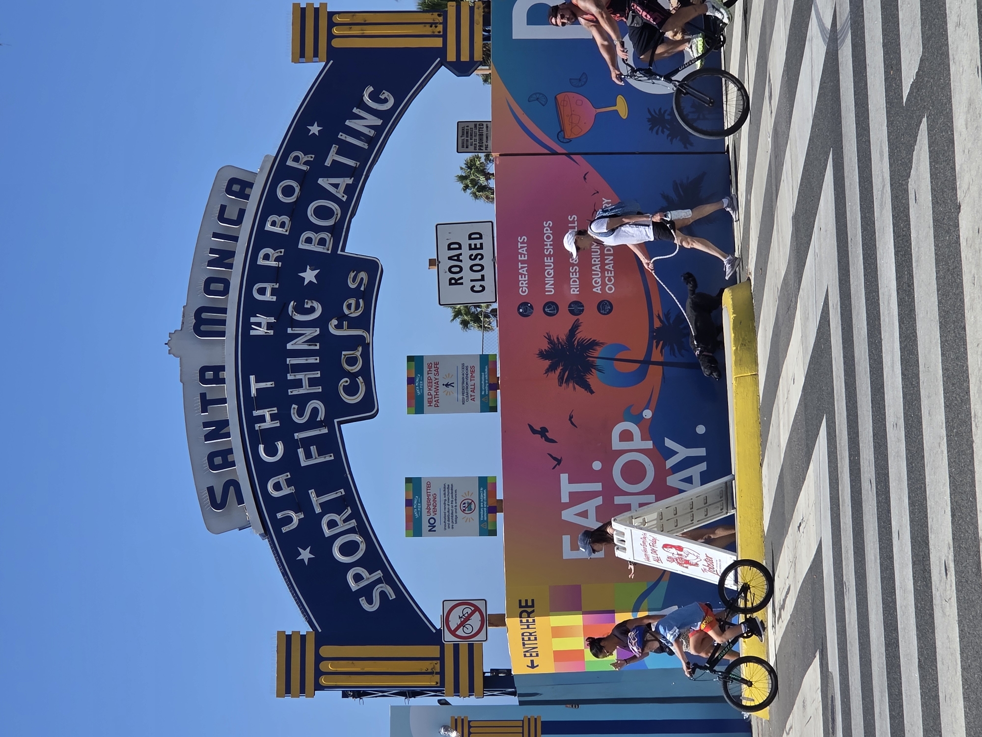 The image captures a view of the Santa Monica Pier entrance in Santa Monica, California, on a sunny day. A large blue archway with white lettering reads "SANTA MONICA YACHT HARBOR SPORT FISHING BOATING CAFES." Below the archway is a colorful mural with palm trees and tropical imagery.
Several people are visible: a young boy rides a bicycle on a crosswalk, a woman walks a black dog on a leash across the same crosswalk, and another person rides a bicycle on the right side of the image. A white A-frame sign stands near the crosswalk, displaying text that includes "Happy Hour Every Day ALL DAY Friday!"
Two official signs are mounted on posts. One indicates "NO UNPERMITTED VENDING," and another instructs "HELP KEEP THIS PATHWAY SAFE" with a pedestrian icon. A "ROAD CLOSED" sign is also present. The environment features a clear blue sky and palm trees in the background. The foreground consists of a black and white striped crosswalk.