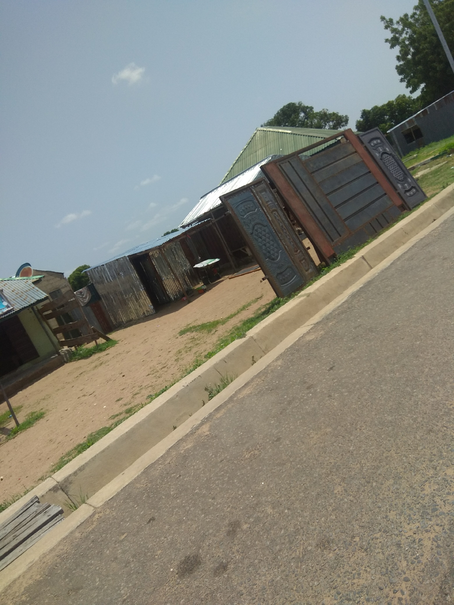 The image depicts a street scene in Jalingo, Nigeria. In the foreground, a paved road runs diagonally from the bottom right to the top left. A concrete curb and a drainage ditch are visible along the edge of the road. Beyond the ditch, a sandy area is present, with some sparse green vegetation and what appears to be a pile of wooden planks. Several corrugated metal structures, likely small shops or dwellings, are situated in the middle ground, some with blue roofs. In the background, a large, decorative metal gate is leaning against some of the structures. Trees and a building with a slanted roof are also visible further in the background. The sky is clear with a few scattered clouds. The scene appears to be an outdoor marketplace or a residential area with commercial establishments.
