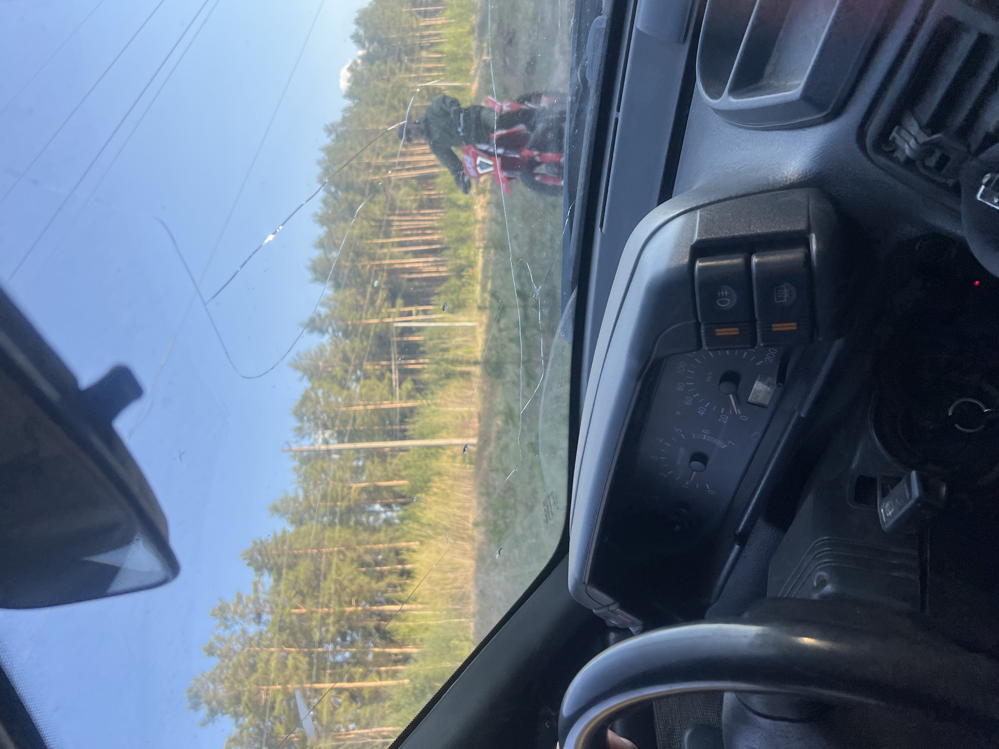 A view from inside a vehicle with a cracked windshield. Outside, a red dirt bike with the number "17" is visible, likely in a rural or wooded area. The vehicle's dashboard is visible, showing a speedometer and other controls. It is not possible to determine the precise location in Chaikovsky, Russia, from the information available in the image.