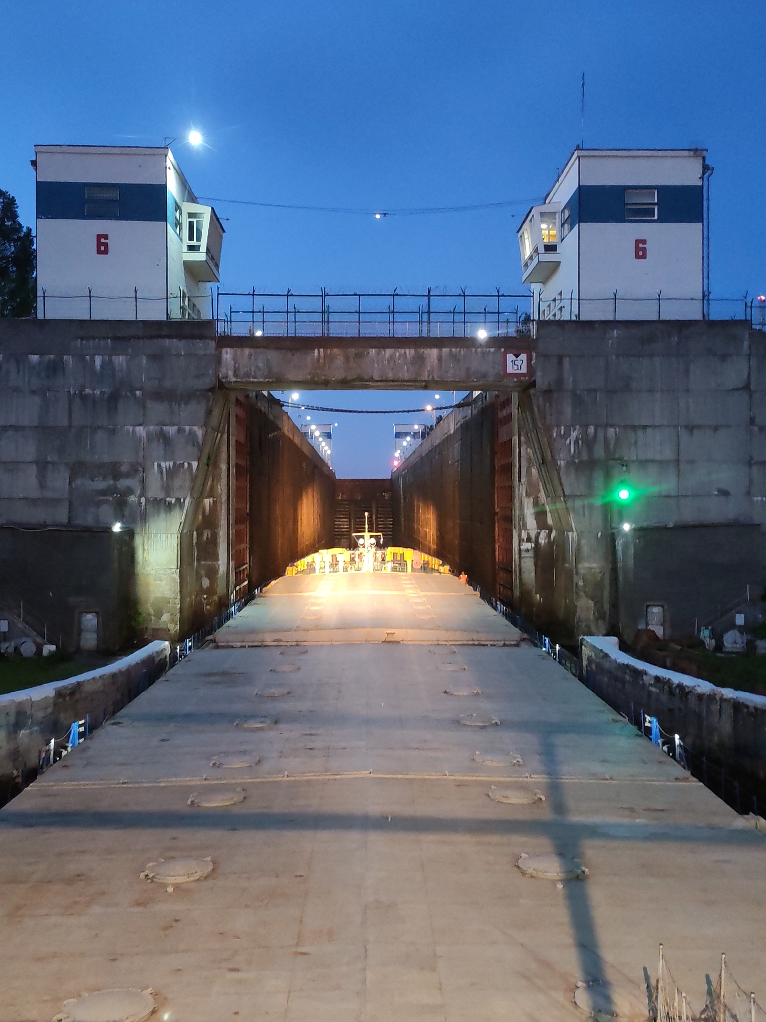The image displays a large ship lock at dusk or night, illuminated by artificial lights, in Devyatiny, Russia. A broad vessel, likely a barge or cargo ship, occupies the foreground, its flat deck with numerous circular hatches extending into the lock chamber.
The lock is defined by imposing, textured grey concrete walls. Perched atop these walls, on either side, are identical control towers. These two-story white buildings feature horizontal blue stripes and the red number "6" prominently on their exterior. Each tower includes windows, some emitting light, and a small cantilevered observation room facing the lock. Antennas are visible on their roofs. A metal fence runs along the top of the lock walls, connecting to the towers.
Bright artificial lights illuminate the interior of the lock and the vessel's deck. A distinct green signal light glows on the right concrete wall of the lock, likely indicating permission or status. Above the lock entrance, a crossbeam displays a red sign with the number "157". The sky is a deep blue, characteristic of twilight, with a bright celestial body, possibly the moon, visible near the left tower. The scene depicts a functional, industrial waterway infrastructure operating during the evening hours.