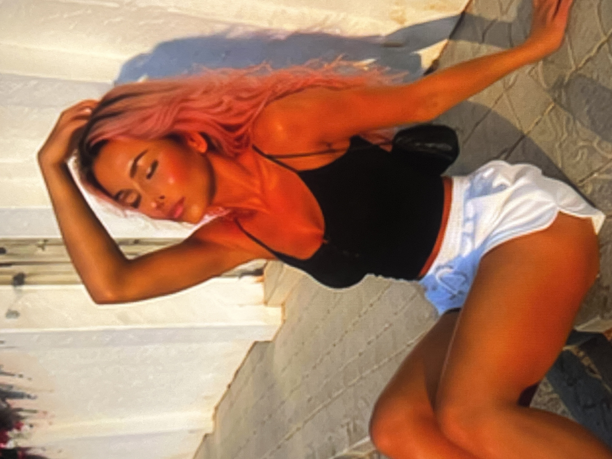 A young woman with pink hair sits on a paved area in front of a white building. She is wearing a black tank top and white shorts. Her left arm is raised and resting on her head, and her eyes are closed. The lighting suggests it is daytime, possibly late afternoon, as evidenced by the long shadows cast on the ground. The overall setting appears to be outdoors, possibly a patio or walkway.