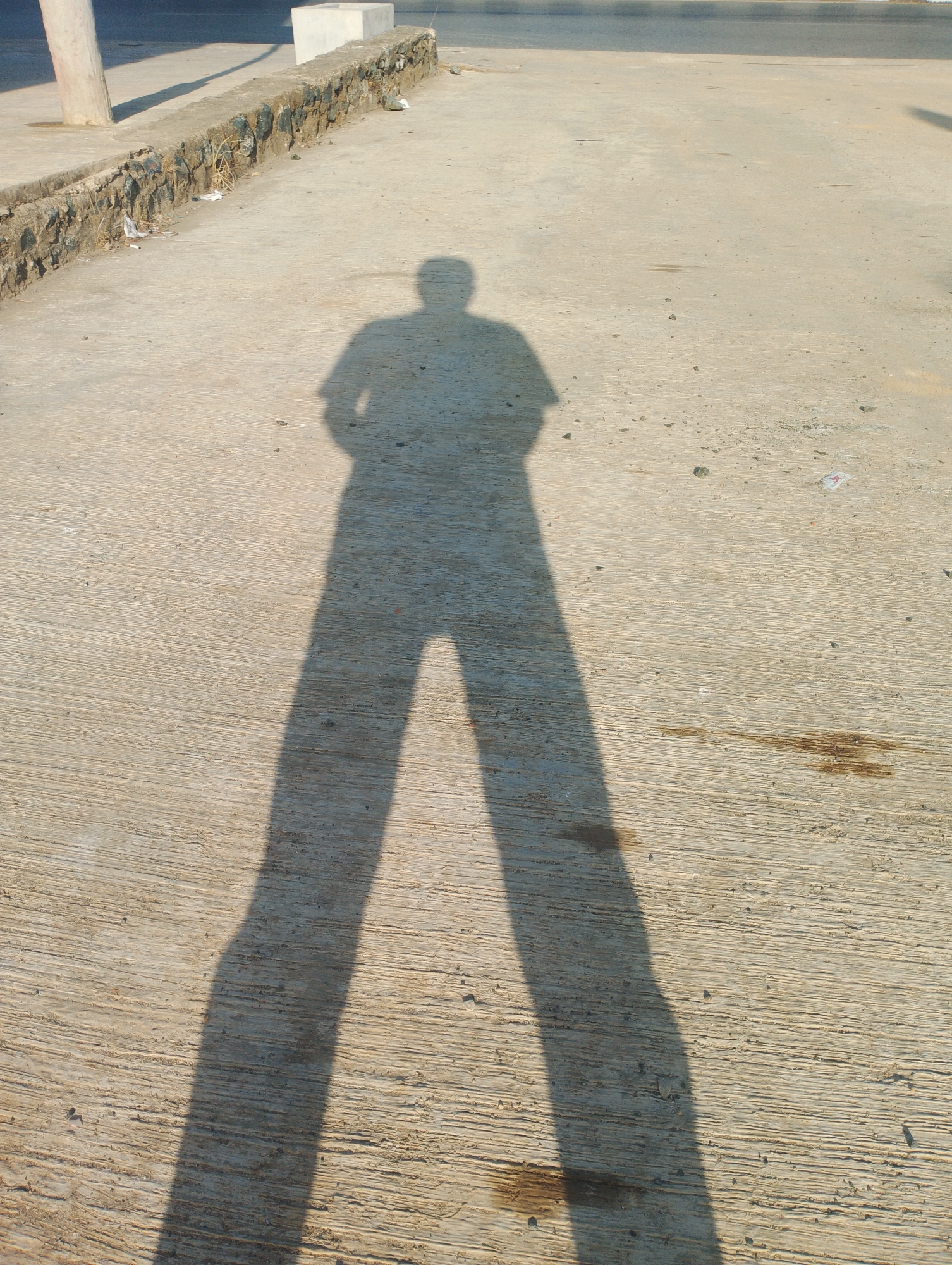 The image captures the elongated shadow of a person standing on a textured concrete surface, likely in Batulicin, Indonesia. The shadow stretches out dramatically, indicating a low angle of sunlight. The ground is a light-colored concrete with visible parallel lines, suggesting a path or pavement. In the background, a low stone wall with rough-hewn stones borders the concrete area. Beyond the wall, a road surface is visible, and further back, a light-colored concrete structure, possibly a planter or a boundary marker, stands out. The overall scene is outdoors, under bright sunlight, casting a sharp shadow and highlighting the texture of the ground. Small debris, including what appears to be discarded paper or plastic, is scattered on the ground near the stone wall.