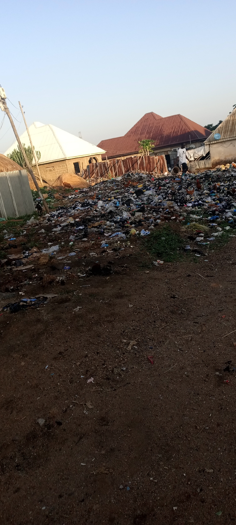 The image shows a large, illegal dumpsite in Jos, Nigeria. The dumpsite is filled with various types of waste, including plastic, paper, and organic material. A man wearing a white shirt and dark pants walks atop the trash pile with buckets. Several chickens are pecking through the refuse. The surrounding environment consists of residential buildings with roofs of varying materials, dirt ground, and power lines. A motorcycle is parked nearby.