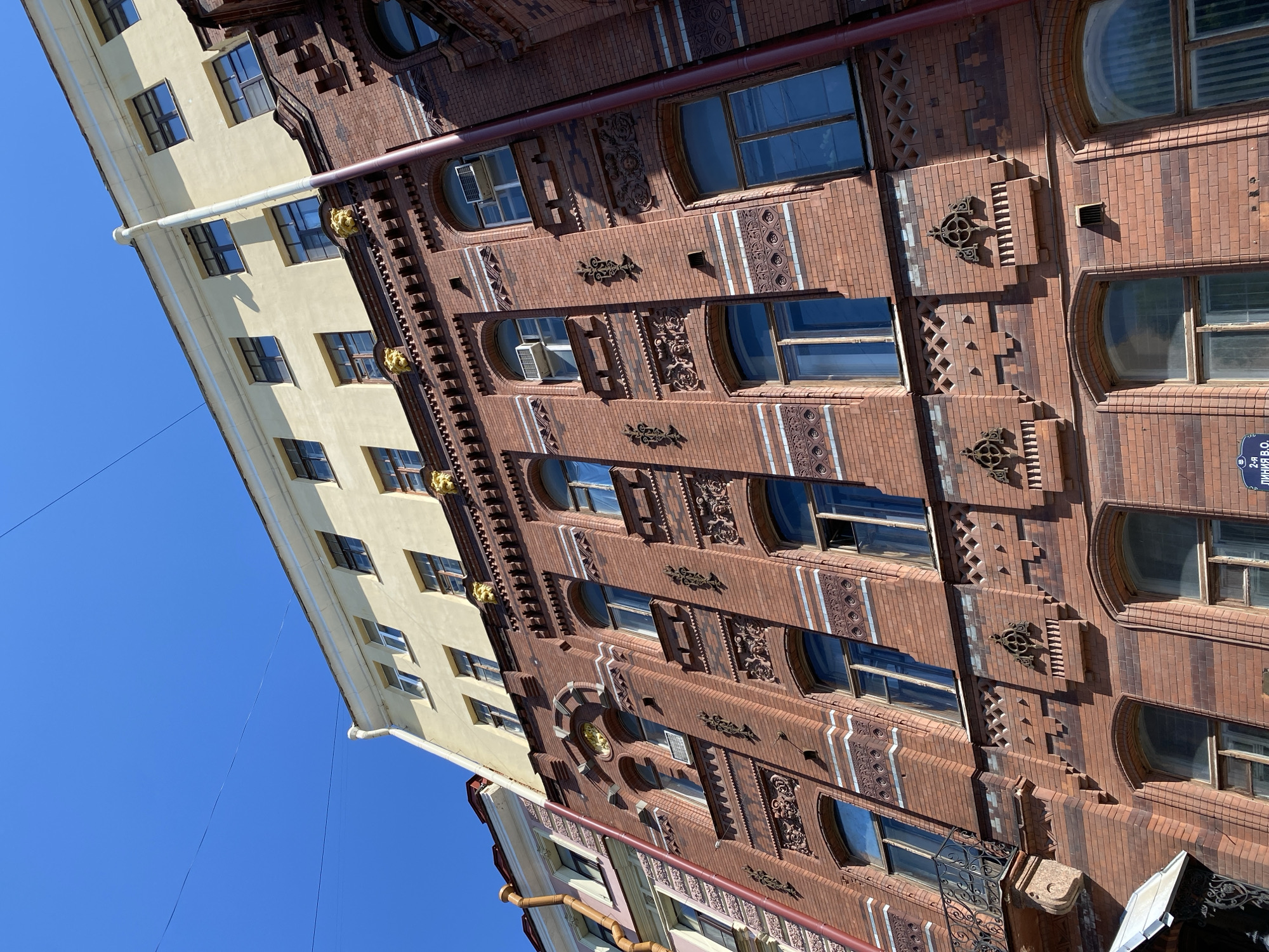 Here's a description of the image:
The image captures a view of two adjacent buildings in what appears to be a European city. The building on the right is predominantly brick, featuring detailed ornamentation, including decorative brick patterns, embellishments around the windows, and architectural features. The windows are rectangular and arched. The building has a sign in Russian, with the word "линия" which might mean "line".
The building on the left has a yellow facade with simple rectangular windows. There are some gilded sculptural elements.
The sky is a clear blue. Overall, the image depicts urban architecture with historical elements.
