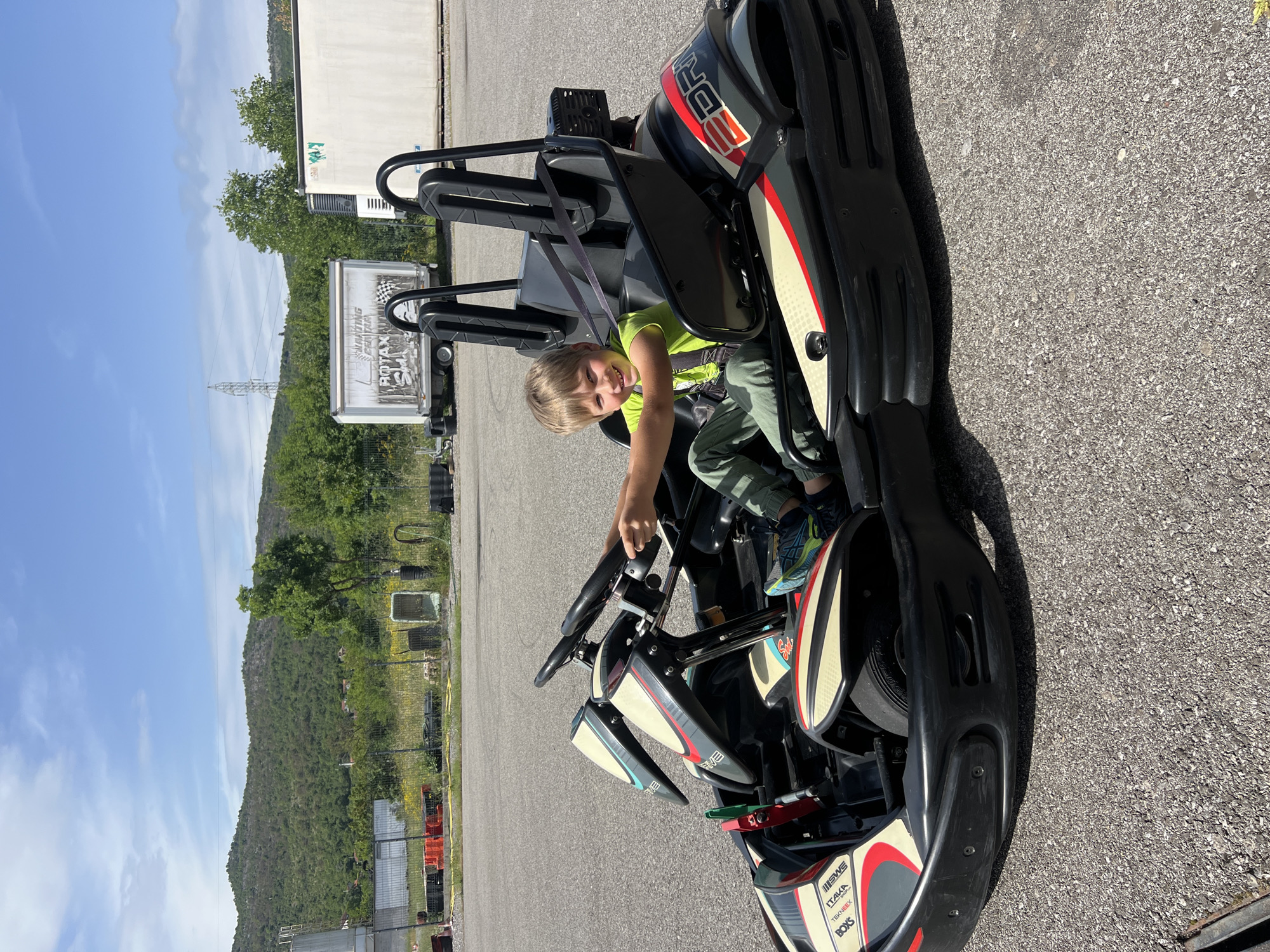 A young child sits in a go-kart on an asphalt surface. The go-kart is black with red and white accents. The child is wearing a green shirt and pants, with a seatbelt secured. In the background is a mountain range, trees, a white building, and a white trailer with writing on the side. The trailer appears to have the word "ROTAX" visible. The location is Bigor, Montenegro.