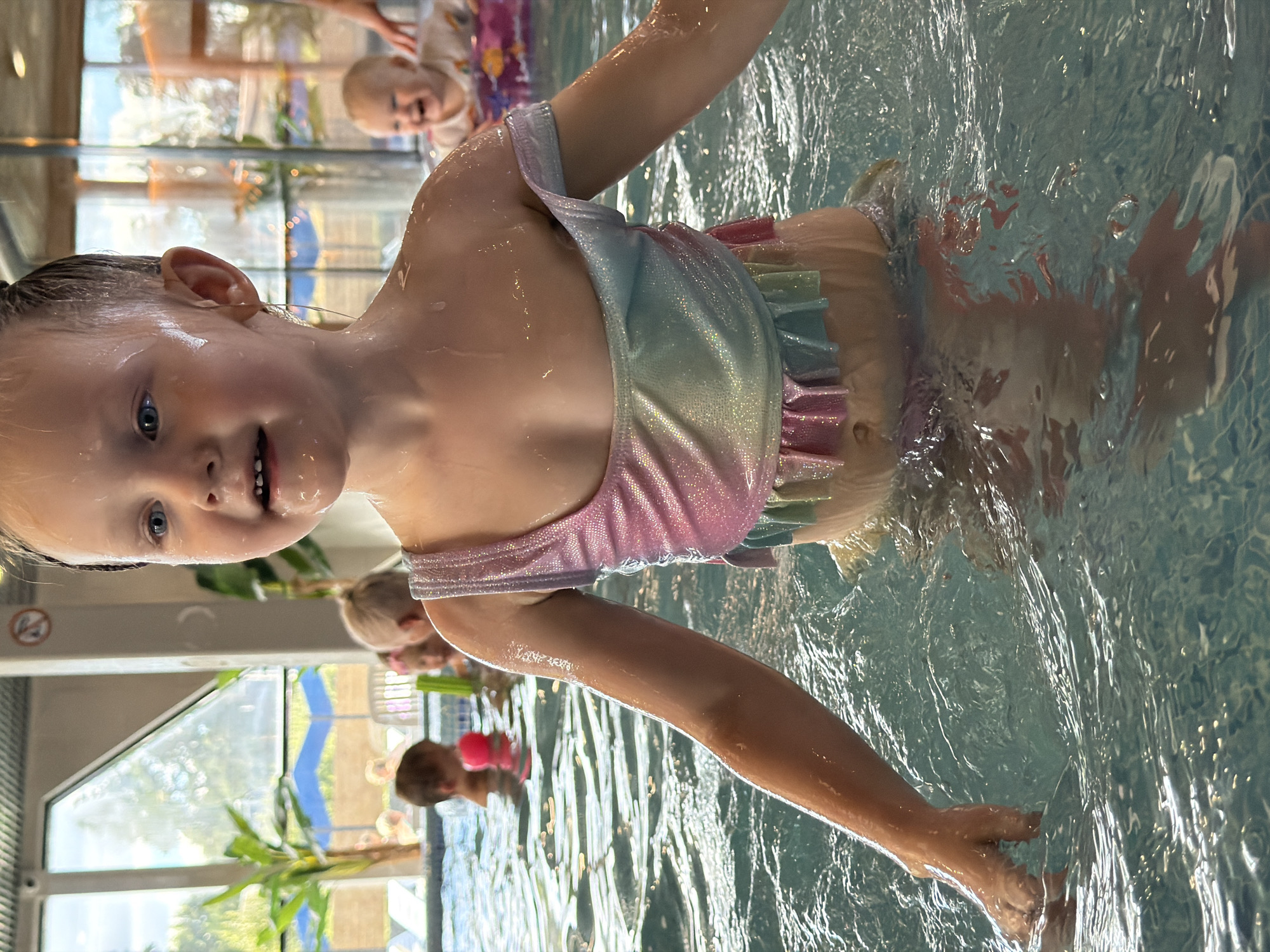 The image depicts a young girl in a swimming pool, her upper body visible above the water. She is wearing a shimmering, iridescent swimsuit with a rainbow color scheme and ruffled trim. Her hair is wet and slicked back, and she has a playful expression with her mouth slightly open. Water droplets are visible on her skin.
In the background, other children are present in the pool. To the left, another child with dark hair is visible, as is a pink floatation device. Further in the background, a baby is sitting in a floating ring and appears to be laughing. The pool area has large windows that offer a view of outdoor scenery with trees and umbrellas. The overall environment suggests an indoor swimming facility.
The location is identified as a swimming pool in the city of Votkinsk, Russia.