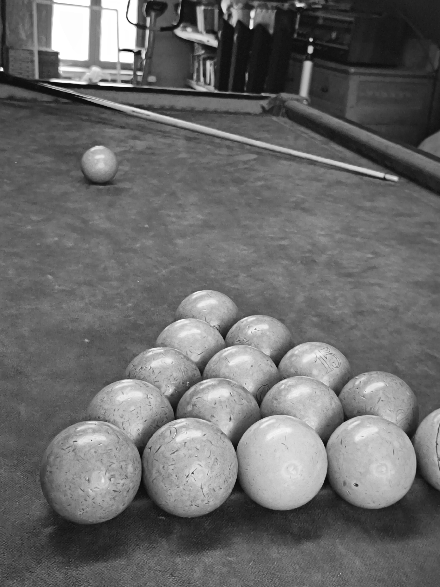 A black and white photograph captures a billiards or pool table in an indoor setting within Терновский округ, Ukraine. Fifteen billiard balls are arranged in a triangular rack formation in the lower-right foreground. These balls exhibit a mottled, worn texture, with one clearly displaying the number "15" and others showing similar faint markings or textures. A single, smoother, lighter-toned cue ball rests independently in the upper-left quadrant of the table's playing surface. A light-colored pool cue lies horizontally across the table, extending from the left rail towards the upper right. The table's felt surface appears textured and well-used, rendered in various shades of gray. The background reveals an interior space, with large, bright windows visible on the upper left, casting light into the room. Indistinct furniture or objects, including what appears to be a wooden cabinet or chest, are visible in the darker background on the right. No individuals are present in the scene.