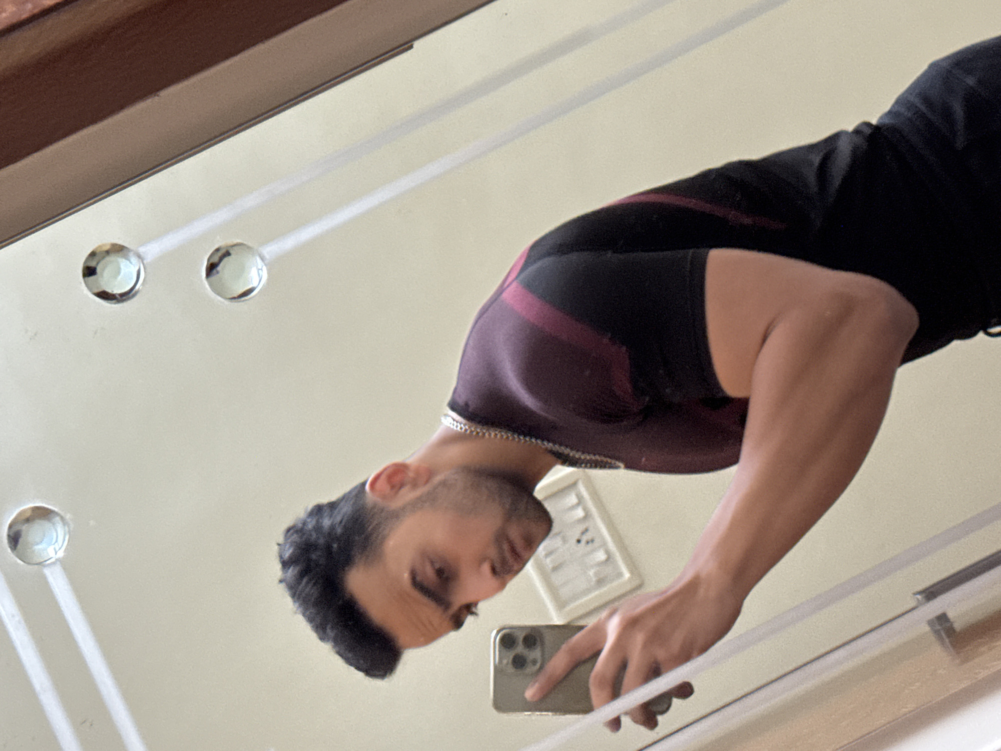 A man stands in front of a mirror, reflected in the image. He is holding a cell phone in his right hand, presumably taking a photo of himself. He is wearing a dark-colored athletic shirt with maroon stripes on the sleeves. The room appears to have light-colored walls and a light-colored mirror frame. There is a small framed object on the wall behind him and decorative mirror attachments. The location is unknown and cannot be determined from the image. The city of Kolkata cannot be confirmed from this image.