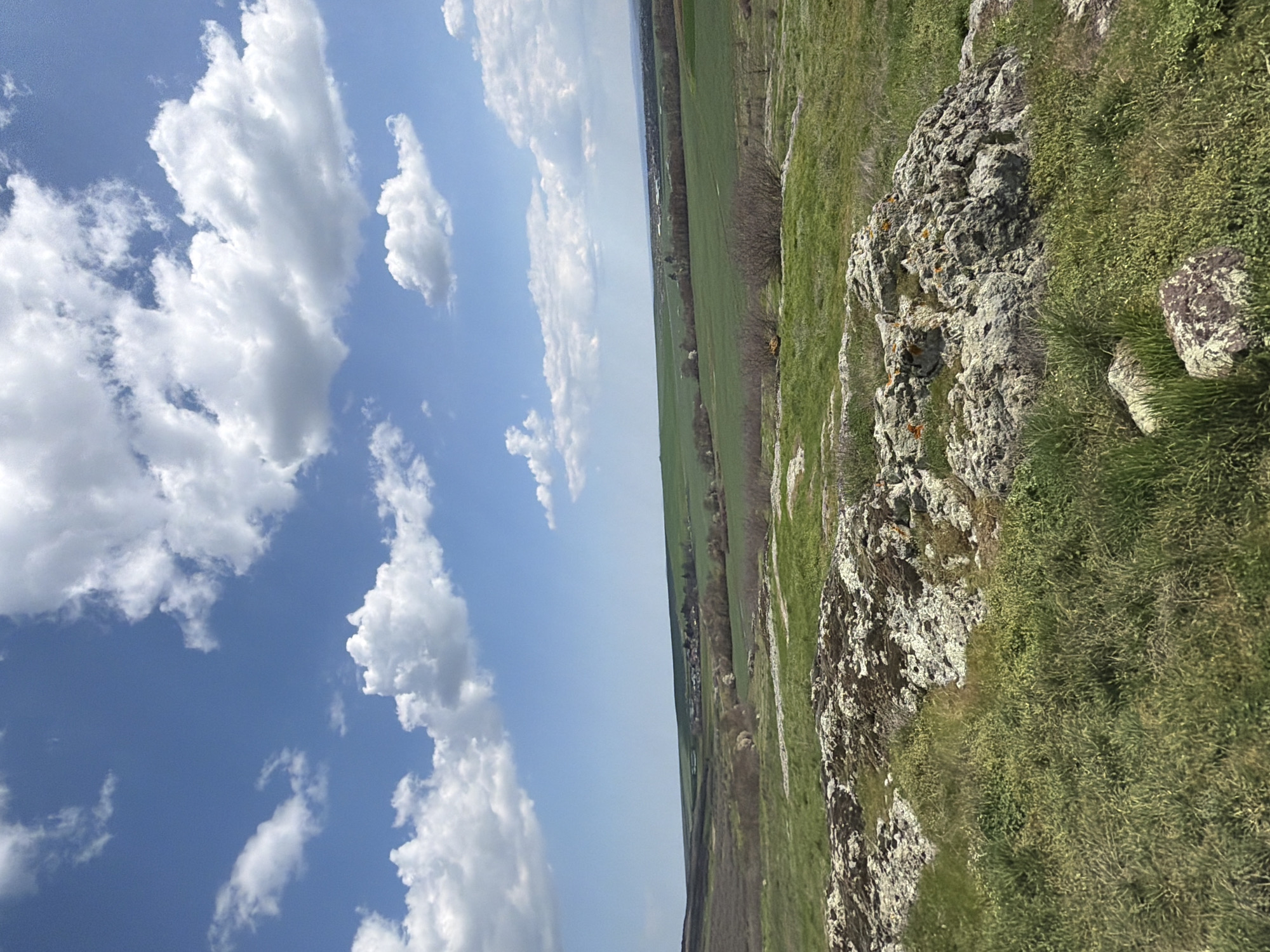 The image depicts a landscape, likely a rural or natural setting, taken in the city of Rusokastro, Bulgaria. The scene is divided roughly in half. On the left side, a blue sky is filled with fluffy white clouds. On the right side, the landscape consists of green fields and grassy terrain dotted with rocks and vegetation. In the distance, there is a line of trees or shrubs, which could indicate the edge of a town or village. The image is oriented on its side, with the sky to the left and the land to the right.