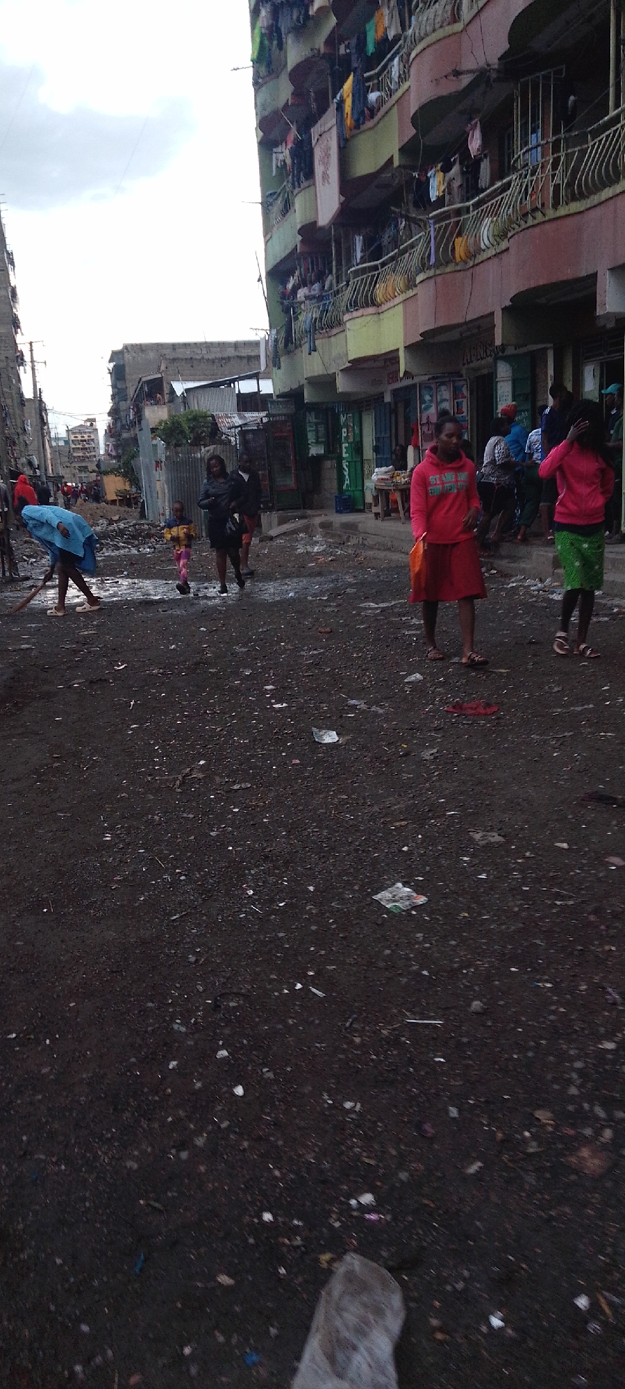 The image depicts a street scene in what appears to be a lower-income area in Nairobi, Kenya. The street is unpaved and covered with litter and debris. Buildings, some with multiple stories and balconies, line the street. Laundry is hanging from some of the balconies. Several individuals are present, including women and children. One person is sweeping or cleaning the street. Other people are walking along the street. There are businesses and shops visible along the street. The sky is overcast.