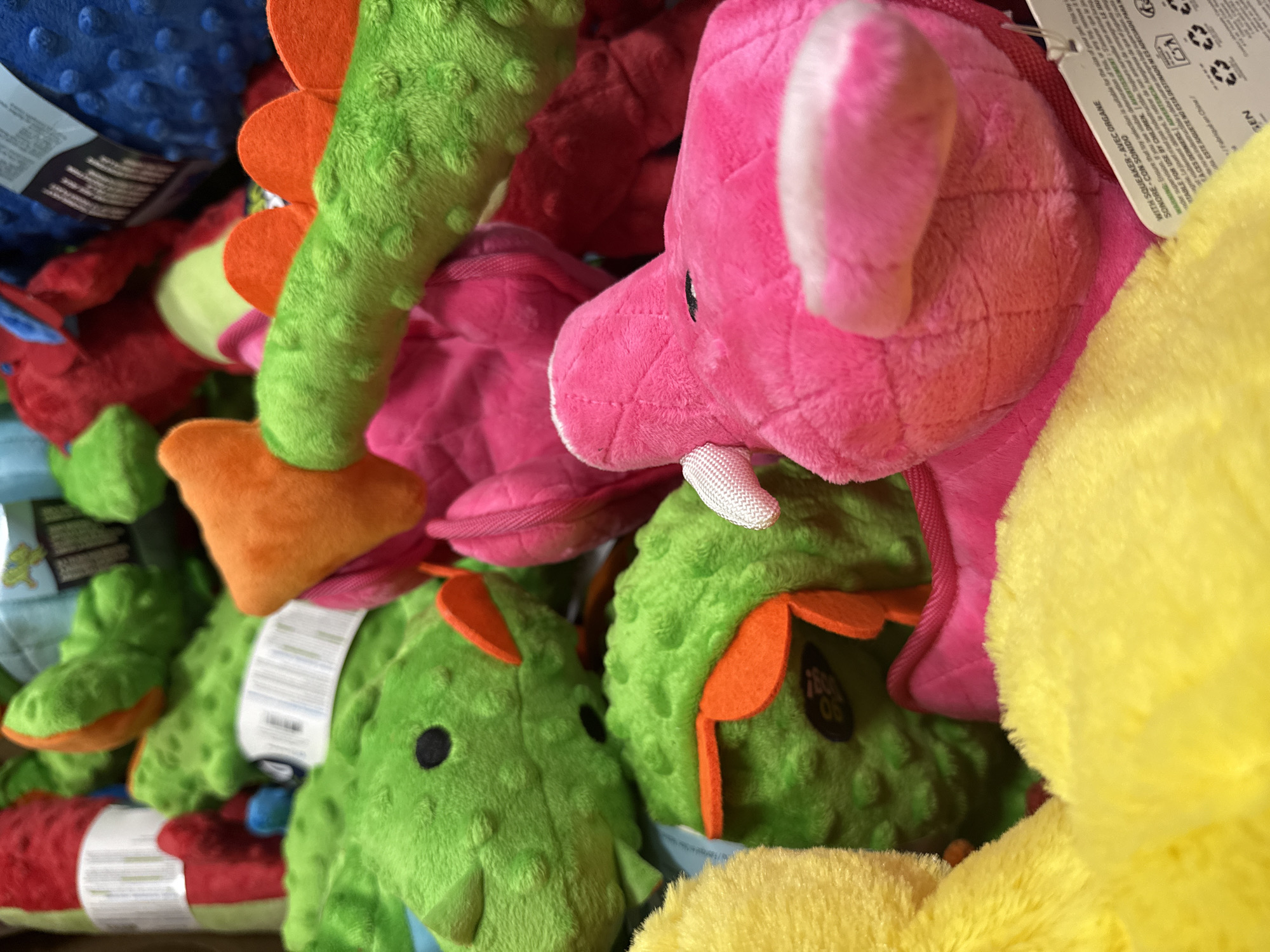 I cannot determine the specific location of this image. The image shows a pile of colorful stuffed animal toys, including green dinosaur figures with orange spines, a pink elephant, a blue figure, a red figure, and a yellow figure. The toys are densely packed, with some having tags still attached. Some of the toys have stitched eyes and rounded shapes. The background is obscured by the volume of toys.