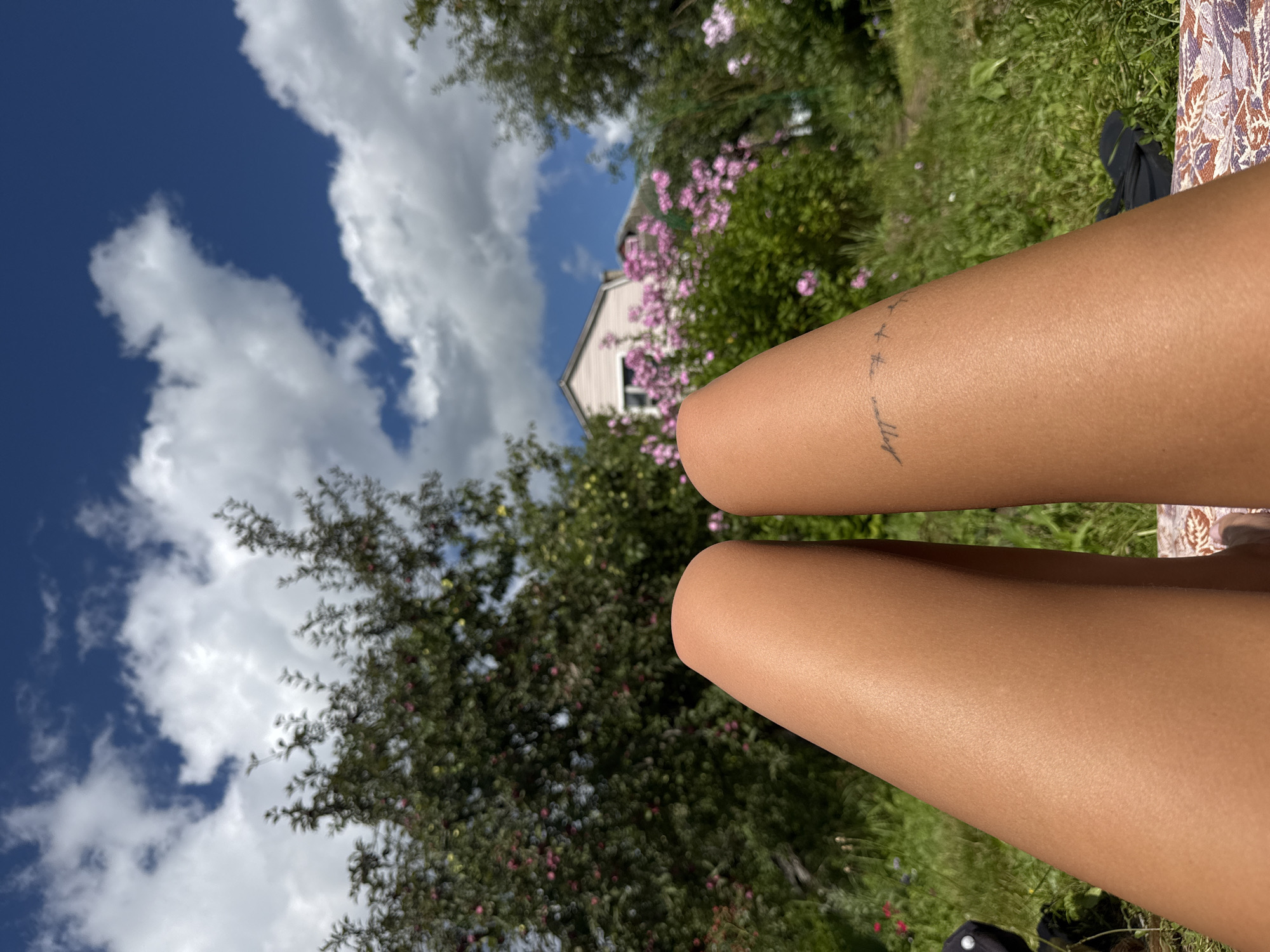 The image shows a first-person perspective of a person lying down outdoors, with their legs prominently featured in the foreground. The legs are tanned and appear to be positioned on a patterned blanket or mat. A faint tattoo of what appears to be a word or phrase followed by a series of crosses is visible on the right leg.
The background suggests a rural or suburban setting, possibly a garden or yard. Lush green grass covers the ground. Behind the grass, there are trees, some with visible fruit or blossoms, and flowering pink bushes. A light-colored house with a pitched roof is visible in the distance, partially obscured by the foliage. The sky above is a bright blue, dotted with fluffy white clouds. The overall impression is one of a sunny day spent outdoors in a peaceful, natural environment. The location is stated as Красная Пресня Снт, Russia, indicating a potential dacha or garden cooperative area.
