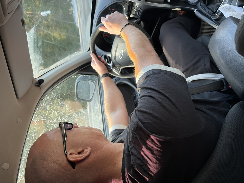 An overhead-view forensic image captures the interior of a vehicle in motion, specifically the driver's position, in Zabegalovo, Russia. The driver is a bald male, viewed from behind his right shoulder, wearing dark sunglasses, a dark short-sleeved t-shirt with light cuffs, and dark trousers. His left wrist bears a dark watch, and his right wrist has a dark beaded bracelet. He is seated in a dark upholstered seat with his seatbelt fastened, and both hands are positioned on the black steering wheel, which features a silver emblem, likely a Chery logo. The dashboard and a portion of the instrument cluster with red needles are visible through the steering wheel. An automatic gear shifter is visible to the driver's right. The windshield and passenger side window are visible, appearing somewhat splattered with debris, possibly road grime or melted snow. Through the windows, a tree-lined, possibly forested, exterior environment is visible, with patches of snow on the ground, indicating a cold or wintery setting. Sunlight illuminates parts of the scene, casting shadows on the driver's shirt and arms. A small black device, possibly a microphone, is attached to the vehicle's interior near the top right of the windshield.