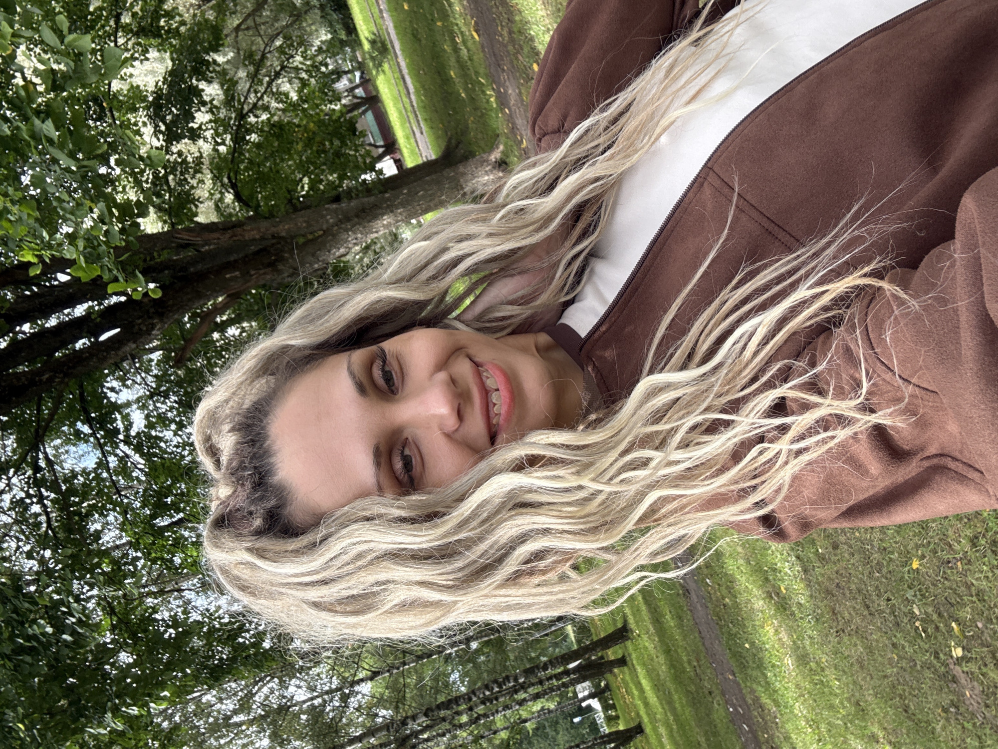 A woman is taking a selfie outdoors in a park. She has long, wavy blonde hair and is smiling. She is wearing a brown zip-up jacket over a white shirt. The background shows trees, grass, and a dirt path. The location is the city of Rusanovo, Russia.