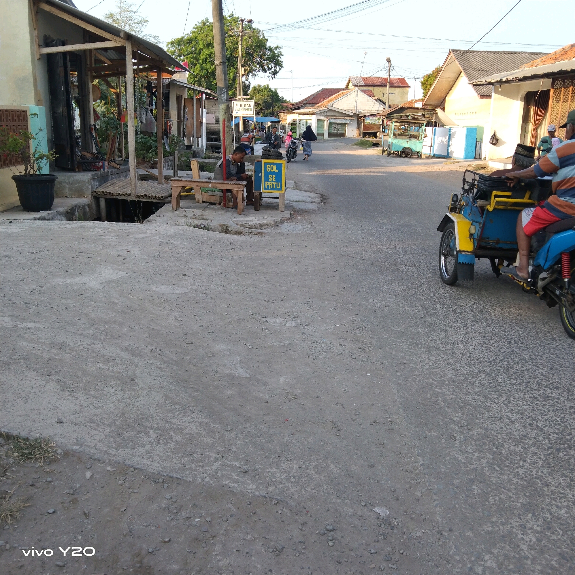 The image depicts a street scene in Karawang, Indonesia. The foreground is dominated by a rough, grey concrete surface, possibly a road or pathway, with scattered small stones and patches of dry grass along the edge. In the mid-ground to the left, a building with a corrugated metal roof and wooden supports is visible. A person is seated at a wooden table in front of the building, looking at a phone. Next to the building, a blue and yellow signage reads "BIDAN DEWI FIRMANSRI." Further down the street, there are other buildings, some with tiled roofs. To the right, a blue and yellow three-wheeled vehicle, resembling a motorcycle with a sidecar, is driving on the paved road. The vehicle has a rider who is partially visible. In the background, more buildings and trees are present under a clear sky. The overall impression is of a modest, possibly rural or semi-urban, street environment during daylight hours.