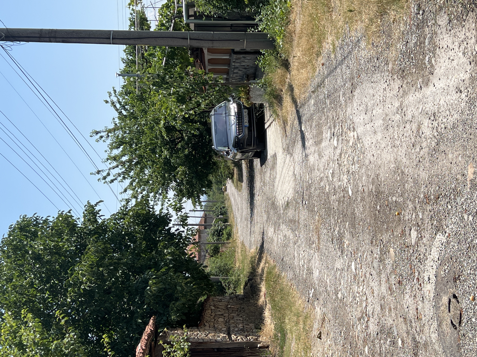 A grey Skoda Karoq is parked on a dirt road in Akandzhievo, Bulgaria. The car is facing forward and is partially obscured by trees. The road is lined with stone walls and houses, and there are power lines overhead. The sky is clear and blue. The car's license plate reads "CB 2355 AE".