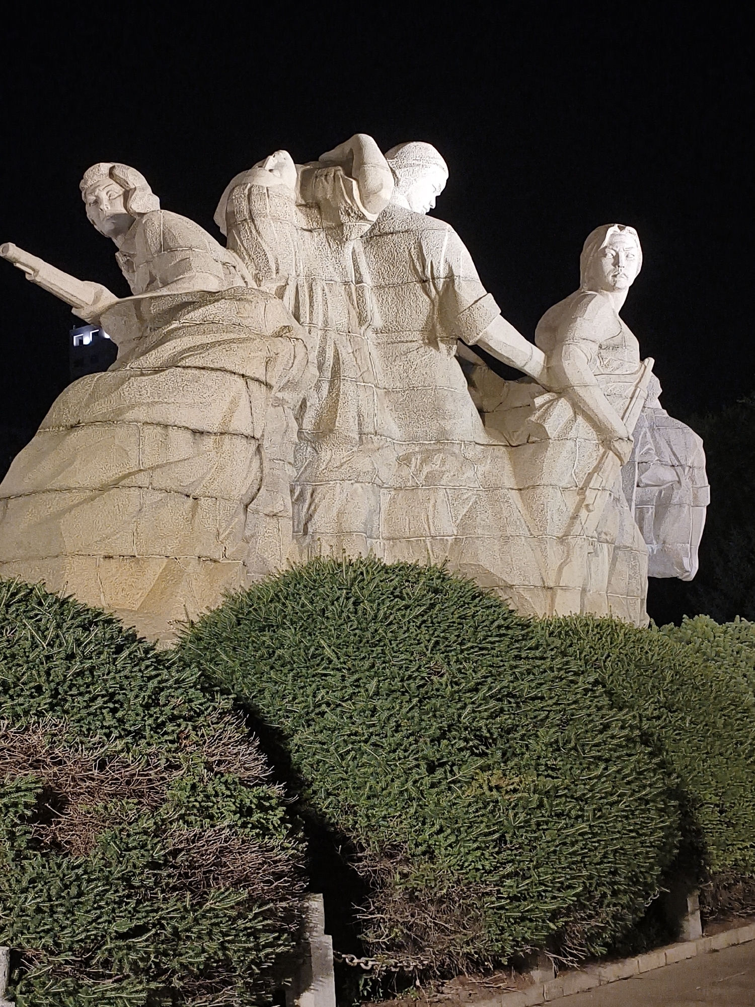 The image shows a nighttime view of a stone sculpture depicting three figures, likely women, dressed in traditional clothing. The figures are carved in a stylized manner, with rough textures and distinct segmented sections. The sculpture is lit from below, casting shadows and highlighting its architectural form. In the foreground, dense green bushes with some dry branches are visible. The background is dark, indicating a night scene, with a faint outline of a building on the left.
This sculpture is located at the Shaanxi History Museum in Xi'an, China. It is an allegorical representation of the "Women's Contingent of the Yellow River," a famous song and dance epic that celebrates the role of women in defending the Yellow River region. The figures in the sculpture are depicted in dynamic poses, suggesting strength and determination. The overall composition conveys a sense of solidarity and resilience.