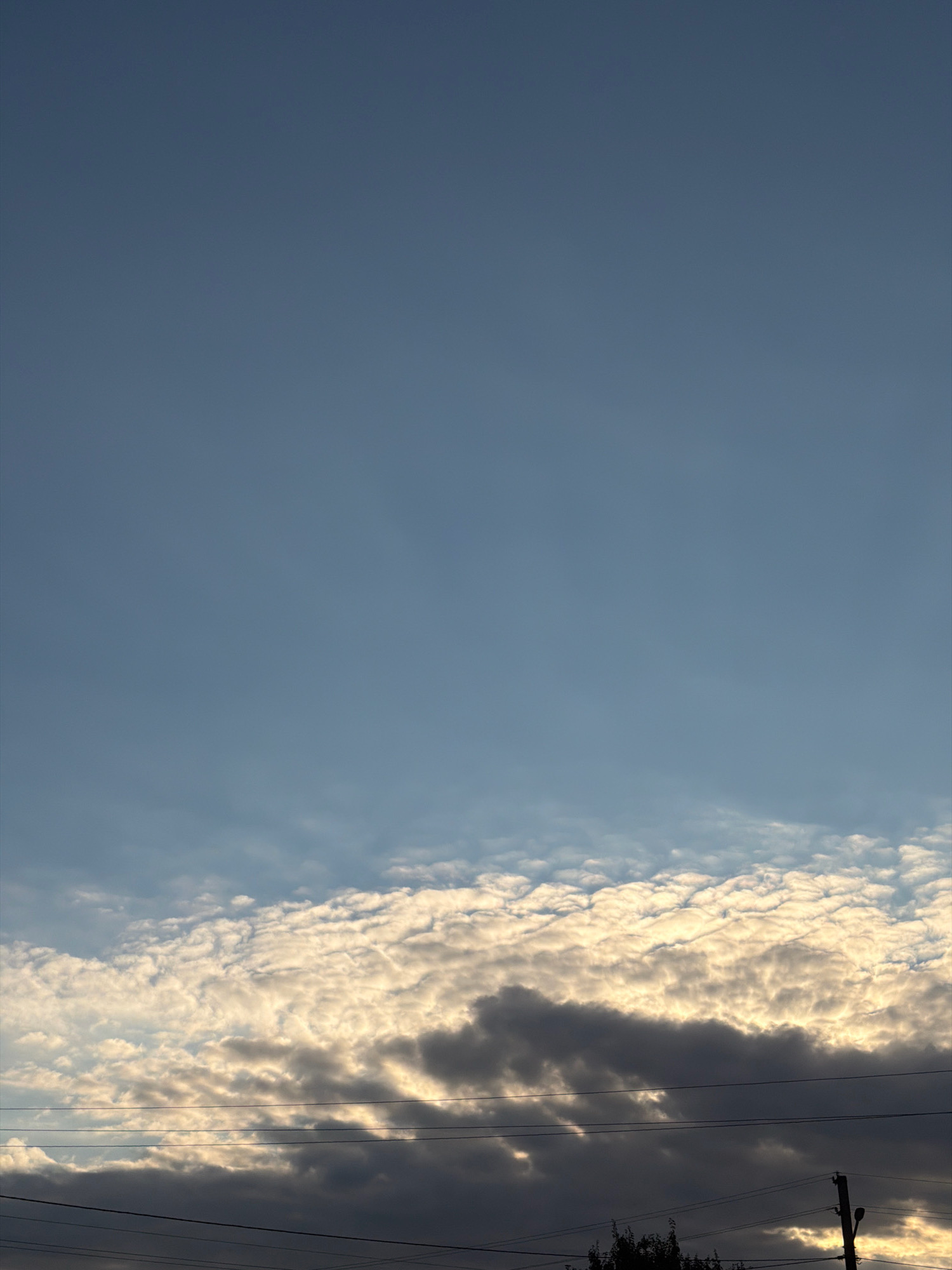 The image shows a sky with a gradient of blue. Towards the bottom of the frame, there are scattered clouds. Some of these clouds are illuminated by the sun, appearing bright white and golden. Other clouds are darker and appear to be in shadow. Horizontal lines, likely power lines, stretch across the lower portion of the image, passing in front of the clouds. A portion of a utility pole is visible on the right side, and the tops of some trees can be seen in the bottom right corner. The scene depicts an outdoor environment, likely in a semi-urban or rural area given the presence of power lines and trees.