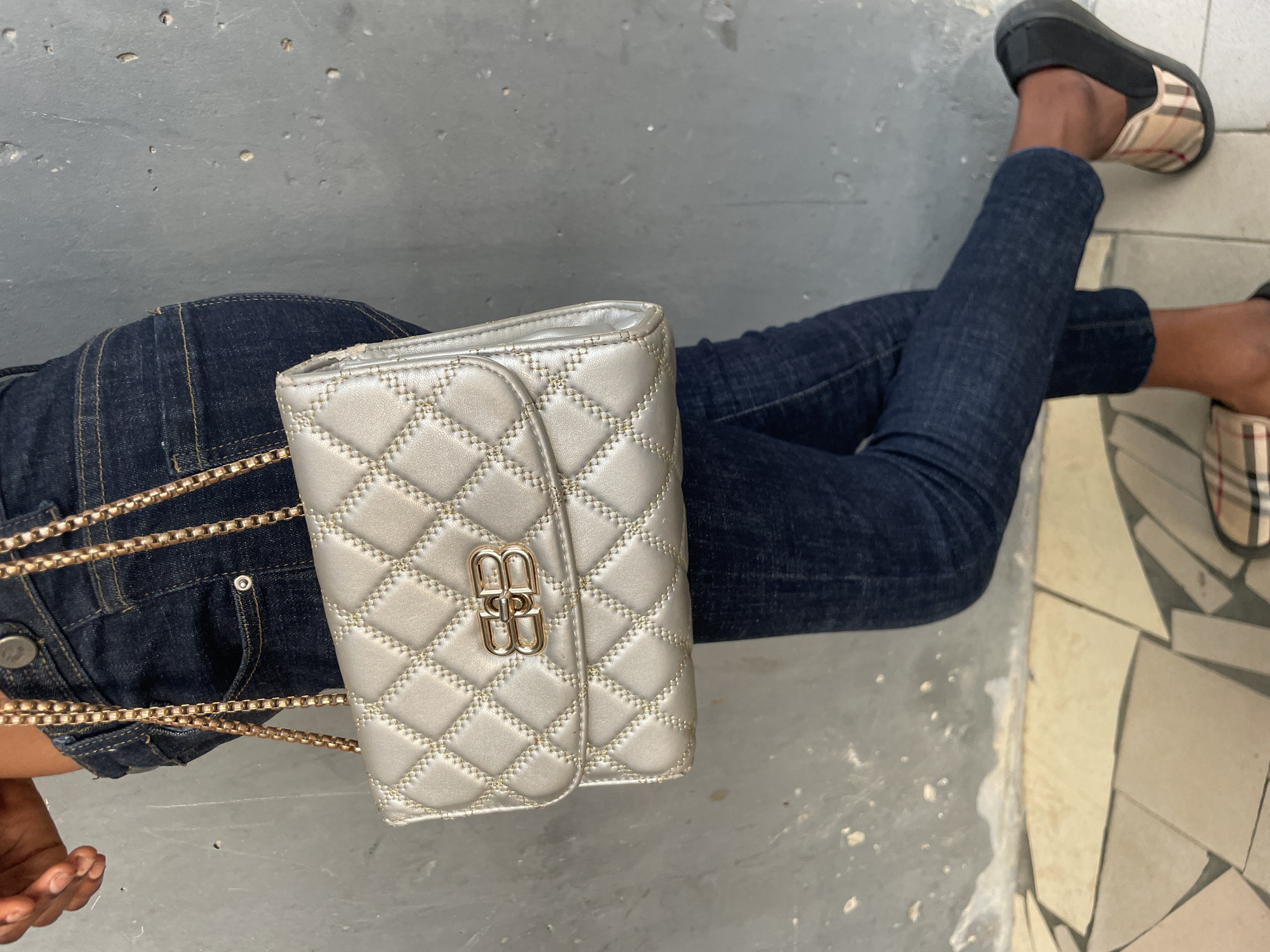 An overhead shot captures the lower body of a person sitting or reclining, featuring dark denim jeans and a prominent silver (or light gold) quilted handbag. The handbag, positioned on the person's lap, has a gold-colored chain strap and a distinct gold clasp with a stylized "BB" logo. The person's dark skin is visible on their hand and feet. They are wearing black slip-on shoes with a beige, red, and black plaid pattern, reminiscent of Burberry.
The scene appears to be outdoors or in a semi-outdoor setting during the daytime, under bright, diffused light. The surface beneath the person is a mix of rough, light gray concrete or tiled flooring and a section of irregularly shaped, light-colored tiles, possibly indicating a pathway, courtyard, or patio. The overall aesthetic is casual, with the handbag as a focal point. Given the context of Badagry, Nigeria, the concrete and tiling could reflect common local urban or residential surfaces. No specific emotions are discernible, and the activity appears to be a posed shot displaying the outfit and accessories.