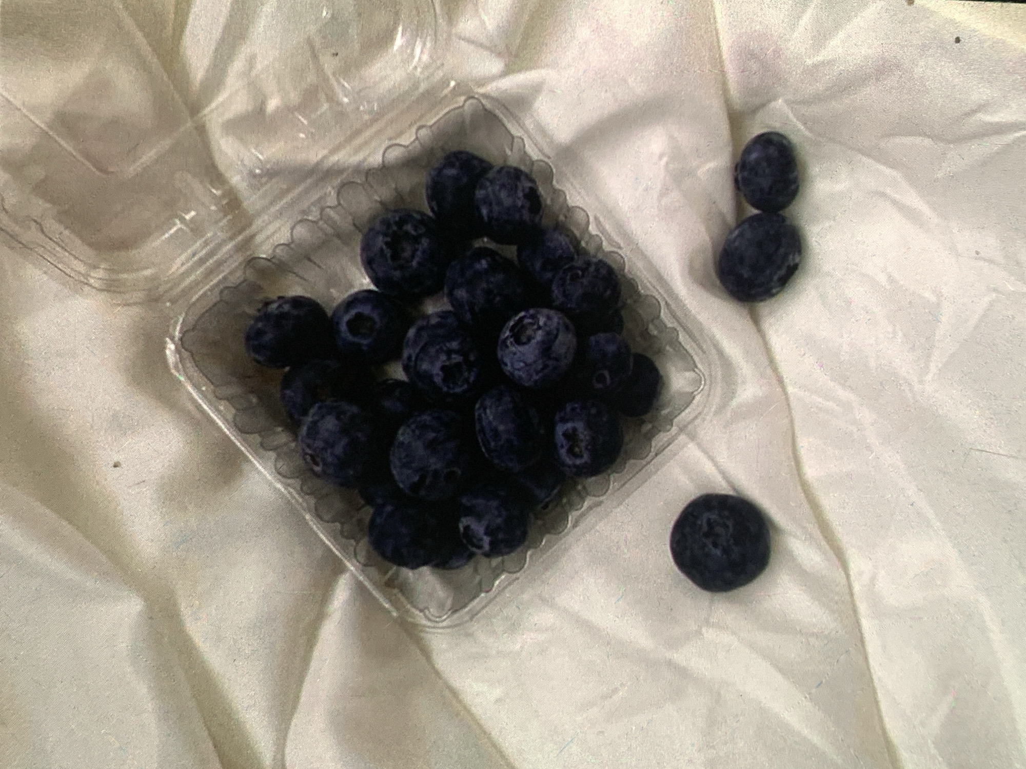 The image shows a clear plastic clamshell container filled with blueberries, placed on a white, textured surface that appears to be fabric. Some blueberries have spilled out of the container and are scattered on the fabric. The lighting is dim, casting soft shadows. There are no people or animals present in the image. The location is not discernible from the visual information provided.