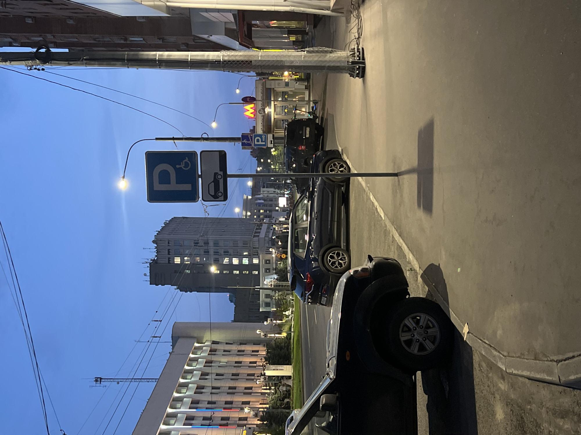 A nighttime street scene in Nizhny Novgorod, Russia, features a blue sky with a gradient from dark blue at the top to a lighter shade near the horizon. Power lines crisscross the sky. On the left, a building with a light-colored facade is illuminated, displaying vertical stripes of red, white, and blue. Further back, a tall, grey, multi-story building with many lit windows is visible. In the foreground, a dark-colored SUV is parked on the left side of the street, its front portion occupying the lower left quadrant of the image.
A sidewalk runs parallel to the street, with a dark blue SUV parked on the right side of the street. In the middle ground, a tall pole supports two traffic signs: a blue square sign with a white "P" and a wheelchair symbol, and below it, a rectangular sign with a white outline of a car. A street lamp on a curved arm extends from the pole, casting a bright light downwards. Further down the sidewalk, there is a metro station entrance with a prominent red "M" logo above the entrance. A scooter is parked near the entrance. The sidewalk is made of concrete. Various electrical poles and wires are present throughout the scene, including a tall metal pole with a complex base in the right foreground. The overall impression is of an urban environment at dusk or early night.