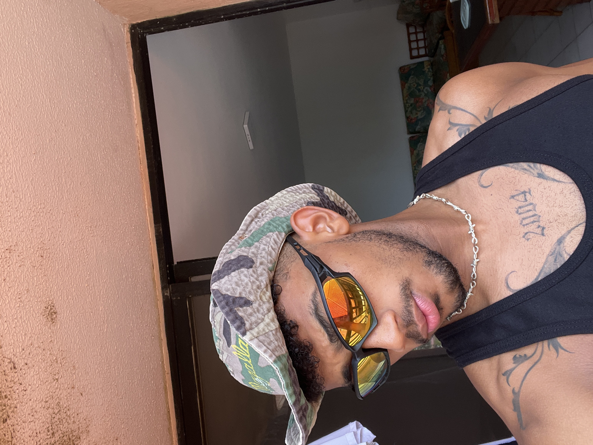 A close-up, selfie-style shot features a young man from the chest up, looking down and to his right. He is wearing a camouflage bucket hat with green and brown patterns, and a silver chain necklace that resembles barbed wire. His black tank top is visible, and he has tattoos on his shoulders and chest, including the number "2004" on his chest. He is also wearing black sunglasses with orange mirrored lenses that reflect buildings. The background shows an interior space with a light-colored ceiling that appears to have some staining or discoloration. A doorway or opening leads to another room where furniture, including sofas and a table, are visible. The scene appears to be indoors, possibly a residential setting. The location is identified as Porlamar, Venezuela.
