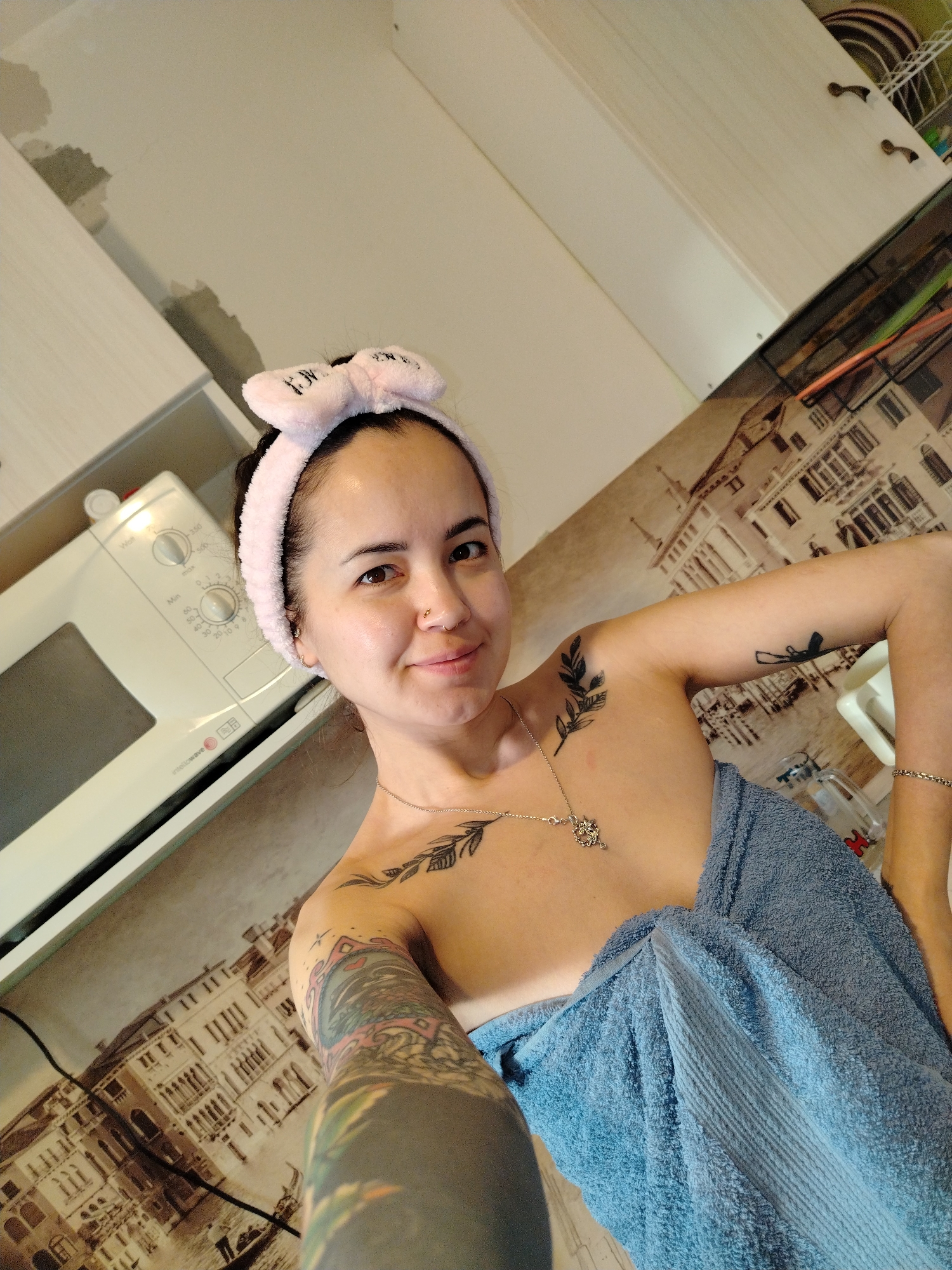A woman takes a selfie indoors, likely within a kitchen in Noviy, Russia. She appears to be in her 20s or 30s, looking directly at the camera with a soft smile. Her dark hair is pulled back, revealing a clean face, and she wears a light pink, plush headband with bunny ears, featuring stylized letters that resemble "F**K" or similar text. She has a small nose piercing and subtle gold hoop earrings.
The woman is wrapped in a light blue terry cloth towel, suggesting she has recently showered or is performing a skincare routine. Around her neck is a delicate silver chain with an intricate pendant. She displays several tattoos: a leafy branch design on her right shoulder, another similar design on her left collarbone, and a prominent, colorful full-sleeve tattoo on her left arm. A small, dark tattoo is also visible on her right bicep, and a gold-colored bracelet adorns her right wrist.
The background indicates a domestic setting. To her left, a white "BOSCH" brand microwave oven is visible, partially recessed into a cabinet. White upper kitchen cabinets are also present. The wall behind her features decorative wallpaper depicting an old European city scene, reminiscent of Venice, with detailed buildings and canals. In the upper left corner of the image, exposed plaster and wall damage suggest ongoing renovation or general wear. The overall scene captures a relaxed and personal moment.