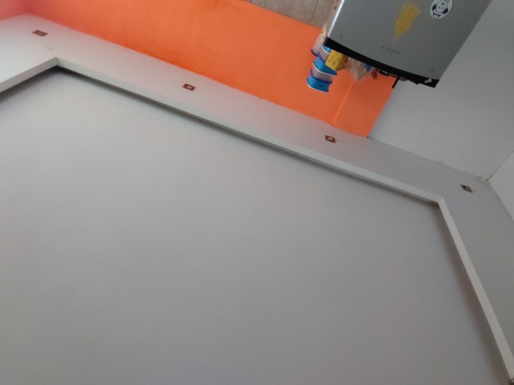 The image presents an interior scene, likely a room or utility space, captured from a tilted perspective. The central focus is a large, unadorned light grey surface, possibly a wall or ceiling. This surface is bordered by a distinct white, L-shaped trim or molding, running across the top and down the left side of the frame. Embedded within this white trim, at spaced intervals, are several small, rectangular, dark brown objects, which could be electrical outlets, mounting points, or recessed lights.
Above the white trim, an orange-painted wall or ceiling section is visible, creating a strong color contrast. Suspended from this orange area is a grey, rectangular appliance, which appears to be mounted overhead. From the underside of this appliance hang several colorful, cylindrical objects, predominantly blue and yellow, resembling stacked containers or filters. The grey appliance features a circular white sticker with a blue graphic, possibly a stylized character or logo, and an irregular yellow sticker. Faint, blurry text may also be present on the appliance below the circular sticker.
The scene suggests a functional area, possibly a kitchen or service space, given the appliance and the clean, uncluttered surfaces. There are no individuals present, and no particular activity is taking place. The lighting suggests it is either daytime or under artificial illumination. No explicit location cues are visible within the image itself, beyond the provided context of Santa Ana, Venezuela.