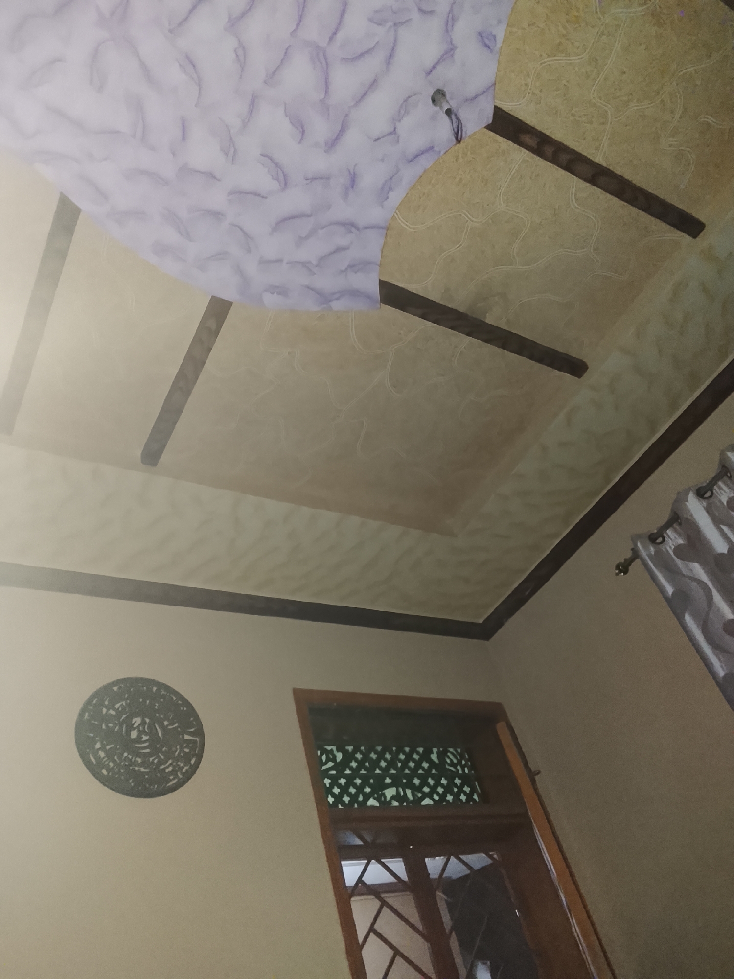 The image displays the interior of a room, captured from a low angle looking upwards and across a wall. The ceiling is a prominent feature, designed with multiple panels. The main section is a light beige or cream color, patterned with subtle, swirling lines. Dark brown wooden beams or strips segment this area. A large, lighter purple or lavender-colored section with a ruffled, fabric-like texture forms a decorative insert, possibly an arch, in the upper left of the ceiling. A small, white light fixture with wires is visible where the purple and beige ceiling sections meet.
The walls are a uniform light beige or cream. On the left wall, a dark, circular decorative piece with intricate, cut-out patterns is affixed. To the right, a wooden-framed window or door is partially visible. It features a dark green screen or grill with a decorative pattern in its upper section, and a grid of wooden cross-bars below. Natural light appears to be entering from this opening, casting a soft glow on the ceiling. A portion of a light-colored curtain, possibly grey or silver with a subtle pattern, hangs on the far right, suspended from a rod with visible rings.
No people are present in the image, and no specific activity or event is depicted. The lighting suggests it is daytime, with natural light entering the room. There is no visible text.