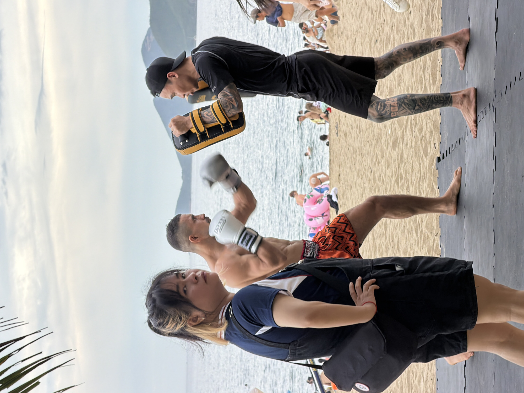 Two men are engaged in a Muay Thai training session on a sandy beach. The man on the left, wearing orange patterned shorts and white boxing gloves, is actively punching. The man on the right, dressed in black shorts and a black t-shirt, wears a black baseball cap and holds a yellow and black padded mitt for the other to strike. He has extensive tattoos on his legs. In the foreground, a woman with dyed blonde-streaked hair, wearing a dark blue shirt with white and red accents and black overalls, stands with her hands on her hips, looking towards the camera. She carries a black shoulder bag. The background shows the ocean, a hazy sky with clouds, and distant mountains, indicative of a coastal city like Nha Trang, Vietnam. Other beachgoers and inflatable rings are visible in the middle ground. The training is taking place on a black mat laid out on the sand.