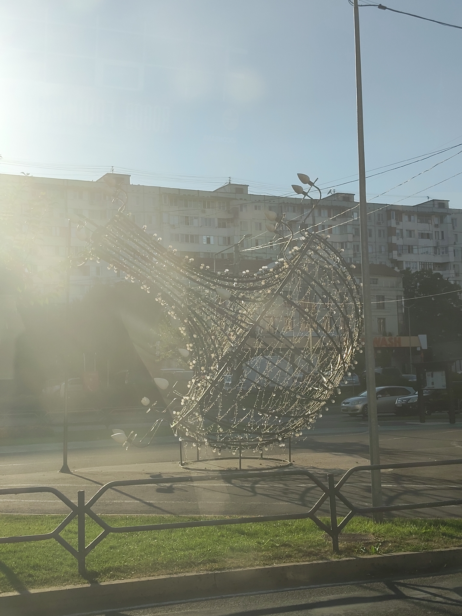An outdoor, sunlit scene captures a large, illuminated decorative sculpture resembling a bulb or seed pod, made of intricate metalwork and tiny lights. The sculpture is positioned in what appears to be a public space, with a road and sidewalk in the foreground, separated by a decorative metal fence. In the background, a large, multi-story apartment building is visible, with a clear sky above. A street lamp stands to the right of the sculpture. Based on the "NASH" sign visible on a building in the background and common architectural styles, this scene is likely located in Chișinău, Moldova. The lighting suggests it is either early morning or late afternoon. No people are clearly visible in the frame.
