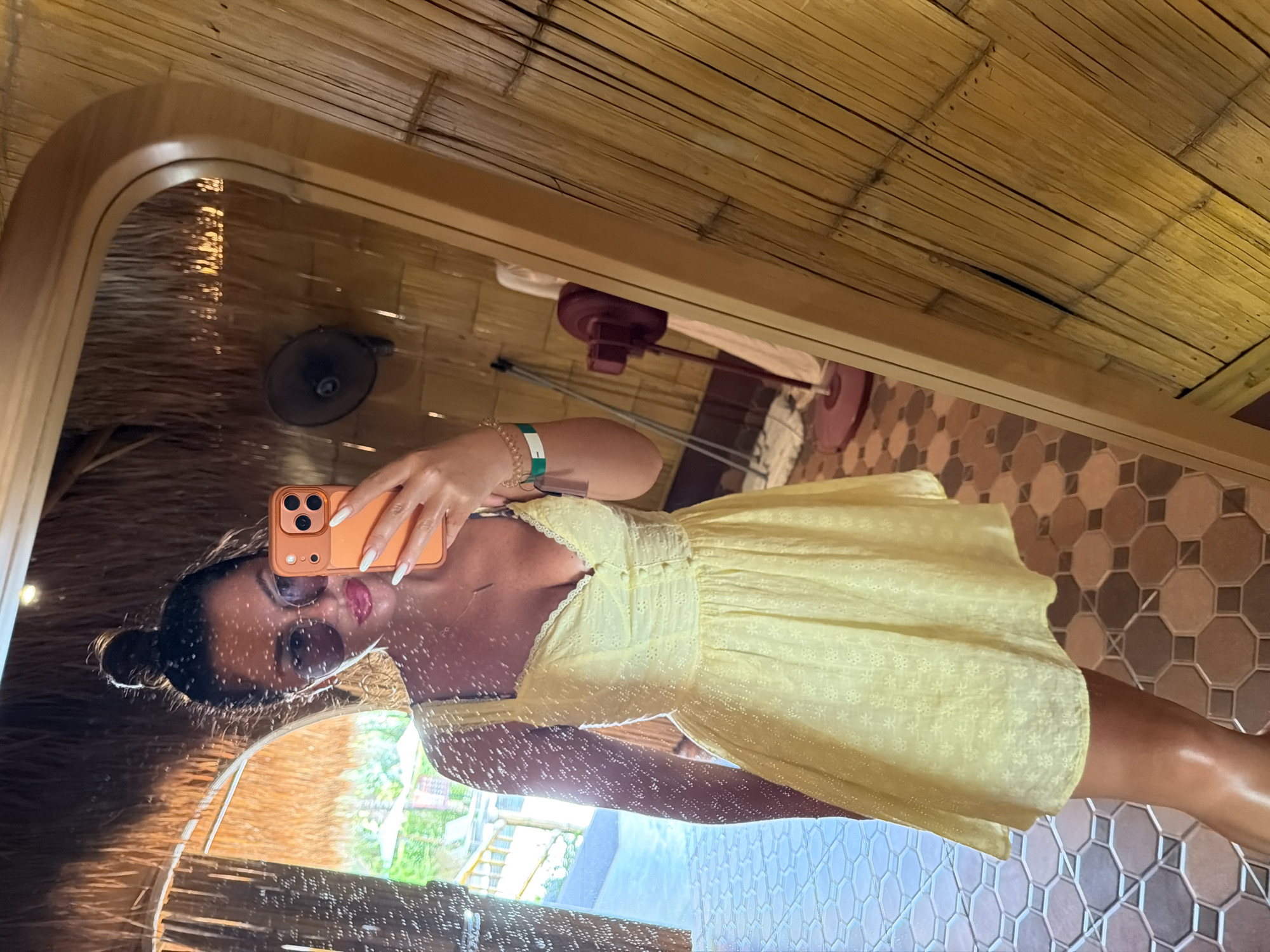A person is taking a mirror selfie in what appears to be a resort or hotel room in Ban Khlong Khian, Thailand. The room has walls made of bamboo and a thatched ceiling. The person is wearing a yellow sundress and has their hair in a bun. They are holding an orange smartphone and wearing sunglasses and jewelry. In the background, a fan and a lamp are visible, along with outdoor scenery reflected in the mirror. The floor is tiled with a geometric pattern.