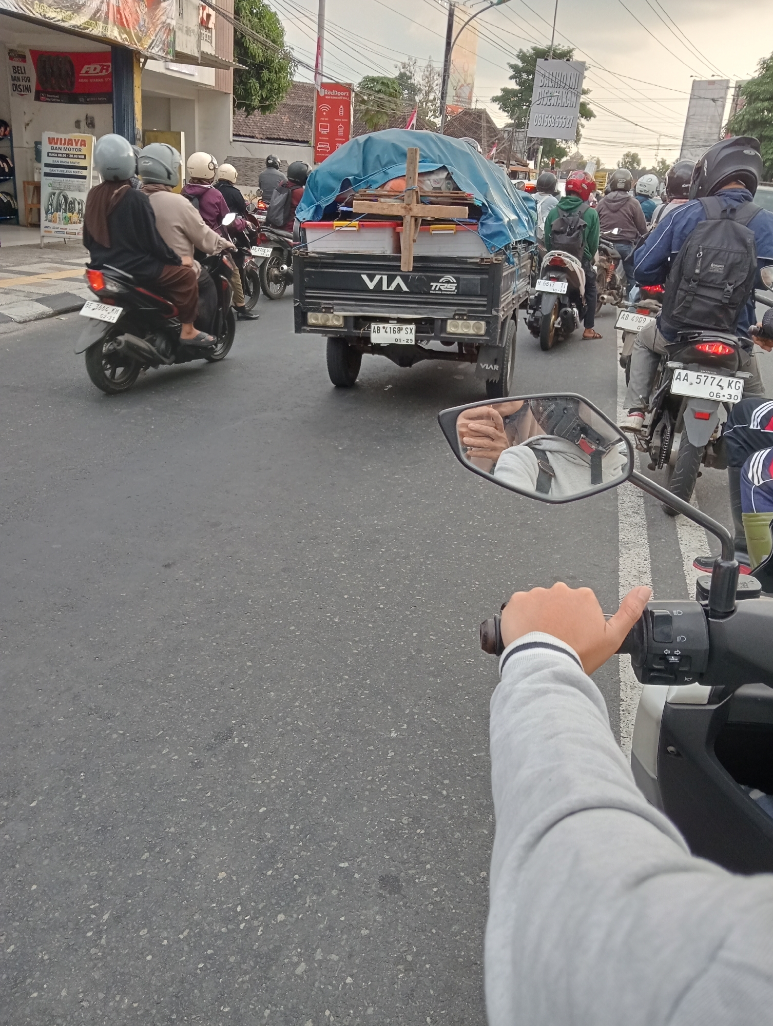 The image depicts a street scene in Sleman, Indonesia, likely during daylight hours with overcast skies. The perspective is from a motorcycle, looking forward and slightly to the right. The foreground shows the rider's right hand on the handlebar and their arm in a grey sweatshirt. A motorcycle mirror reflects the rider's face, wearing a helmet, and the back of another person.
In the mid-ground, traffic is moving on a paved road. Several motorcycles are visible, with riders wearing helmets and varying attire. A prominent object is a small truck, covered with a blue tarp, carrying cargo. The truck has "VIAR" branding. License plates are visible on some vehicles, including one that reads "AA 5774 KG".
The background reveals buildings lining the street, including shops and signs with Indonesian text, such as "WIJAYA BAN MOTOR" and "RedDoorz". Trees and utility poles are also present. The overall impression is of a busy urban or semi-urban environment with moderate traffic flow.