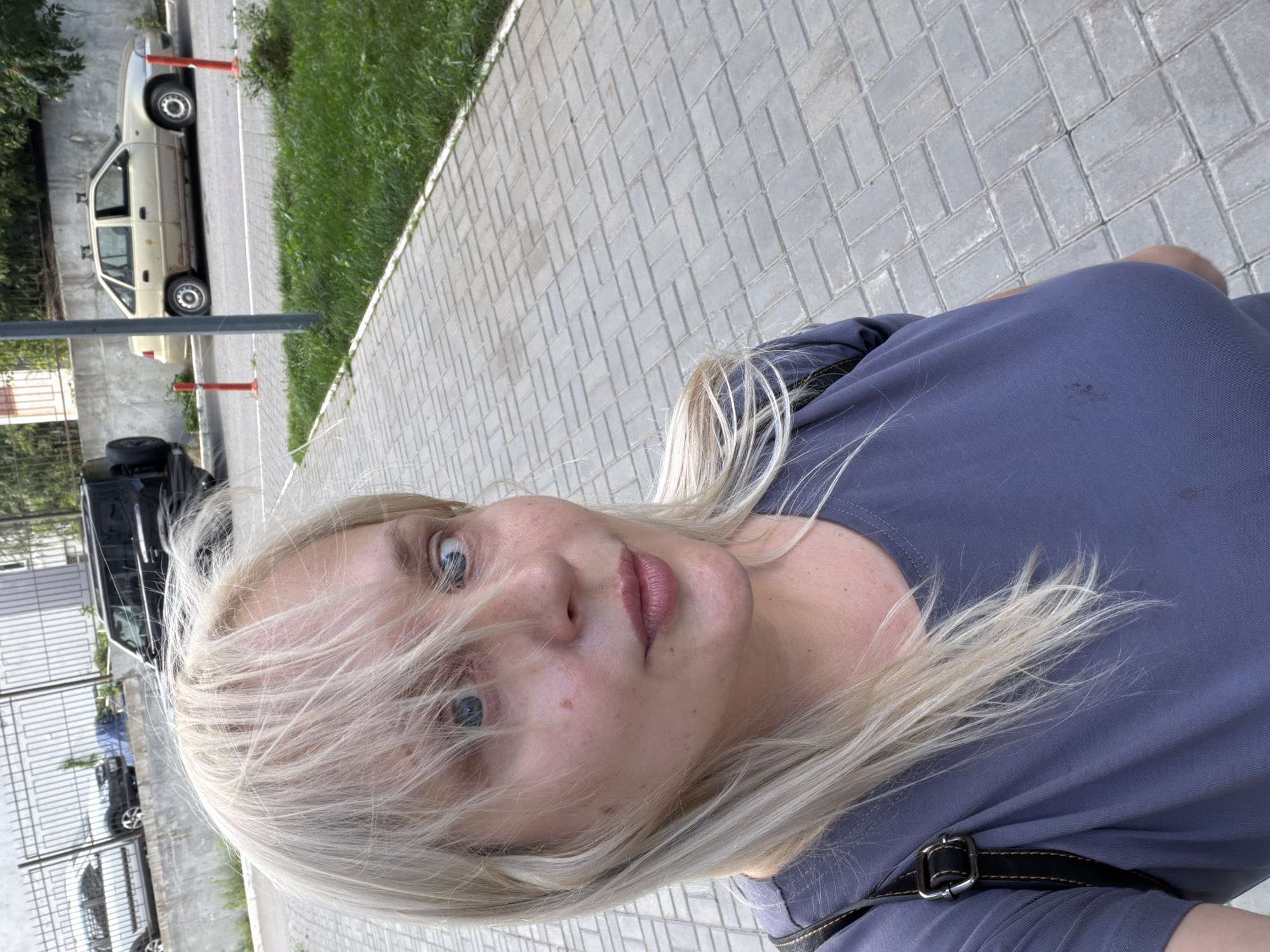 A medium shot selfie shows a Caucasian woman with blonde hair and blue eyes. She is wearing a gray t-shirt and a black backpack. The woman is standing on a paved sidewalk in front of a grassy area. In the background, there are several cars and a metal fence. The scene appears to be in an urban environment. The image was taken outdoors during the day, with natural lighting.