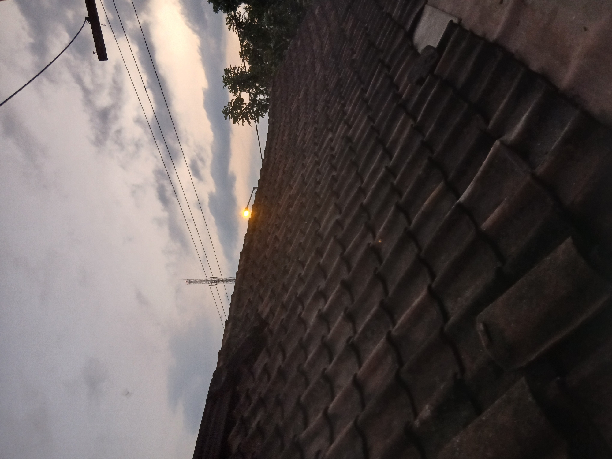 This is a close-up, low-angle shot of a dark, tiled roof under a cloudy sky at dusk. The tiles are oriented diagonally from the bottom left to the top right of the frame. In the upper portion of the image, a telephone pole with wires is visible, and to the right, several trees are silhouetted against the fading light. A street lamp with a bright yellow light is also visible, positioned above the roof and to the left of the trees. The scene appears to be in the city of Tenggarang, Indonesia.