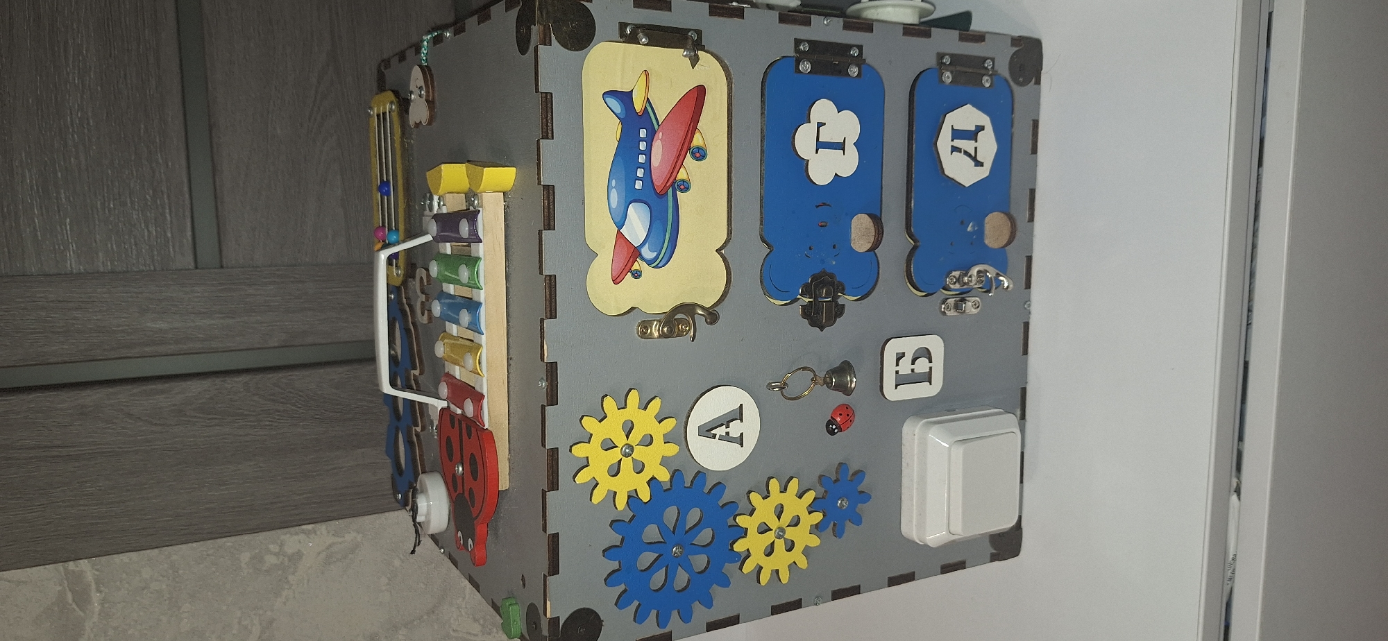 A busy cube toy is displayed on a white surface against a grey wooden wall. The toy has various interactive elements for children, including colorful gears, letters, a xylophone, a ladybug, and different types of latches and locks. On one side of the cube, there is an illustration of a blue airplane. The other sides of the cube feature Ukrainian letters. The toy is positioned to the left of center in the frame. The background wall appears to be a textured wood paneling. The lighting suggests an indoor setting. The exact location within Bila Tserkva, Ukraine cannot be determined from the image.