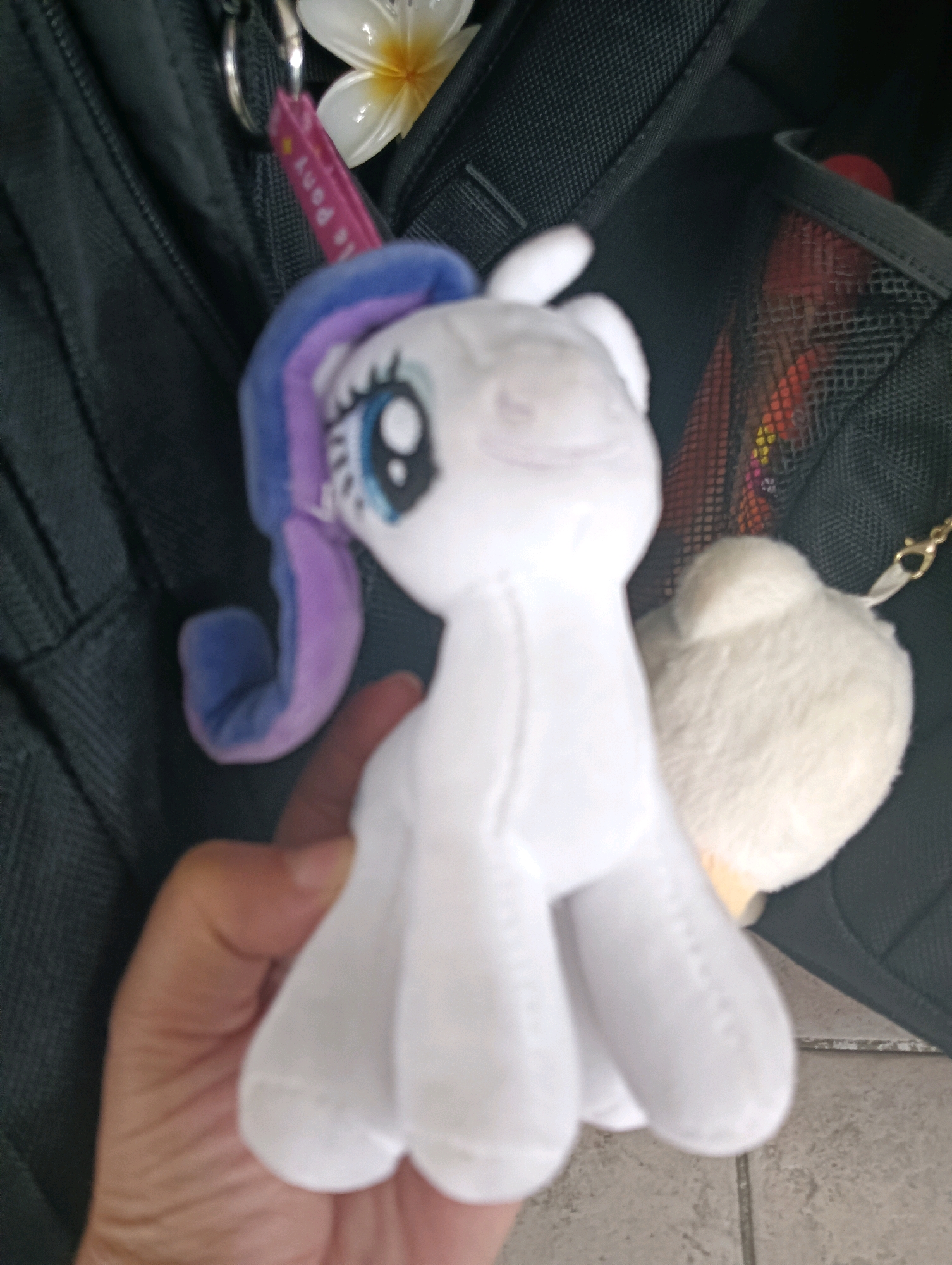 A close-up, first-person perspective shows a hand holding a white stuffed toy unicorn with blue and purple accents. The unicorn has large blue eyes and is partially obscured by the hand. In the background, a black backpack is visible, along with a white flower and a small white fluffy object, possibly another toy or accessory. The scene appears to be outdoors, with a tiled surface and what looks like a street visible in the lower portion of the image. The lighting suggests it is daytime. The location is not identifiable as Tomohon, Indonesia.