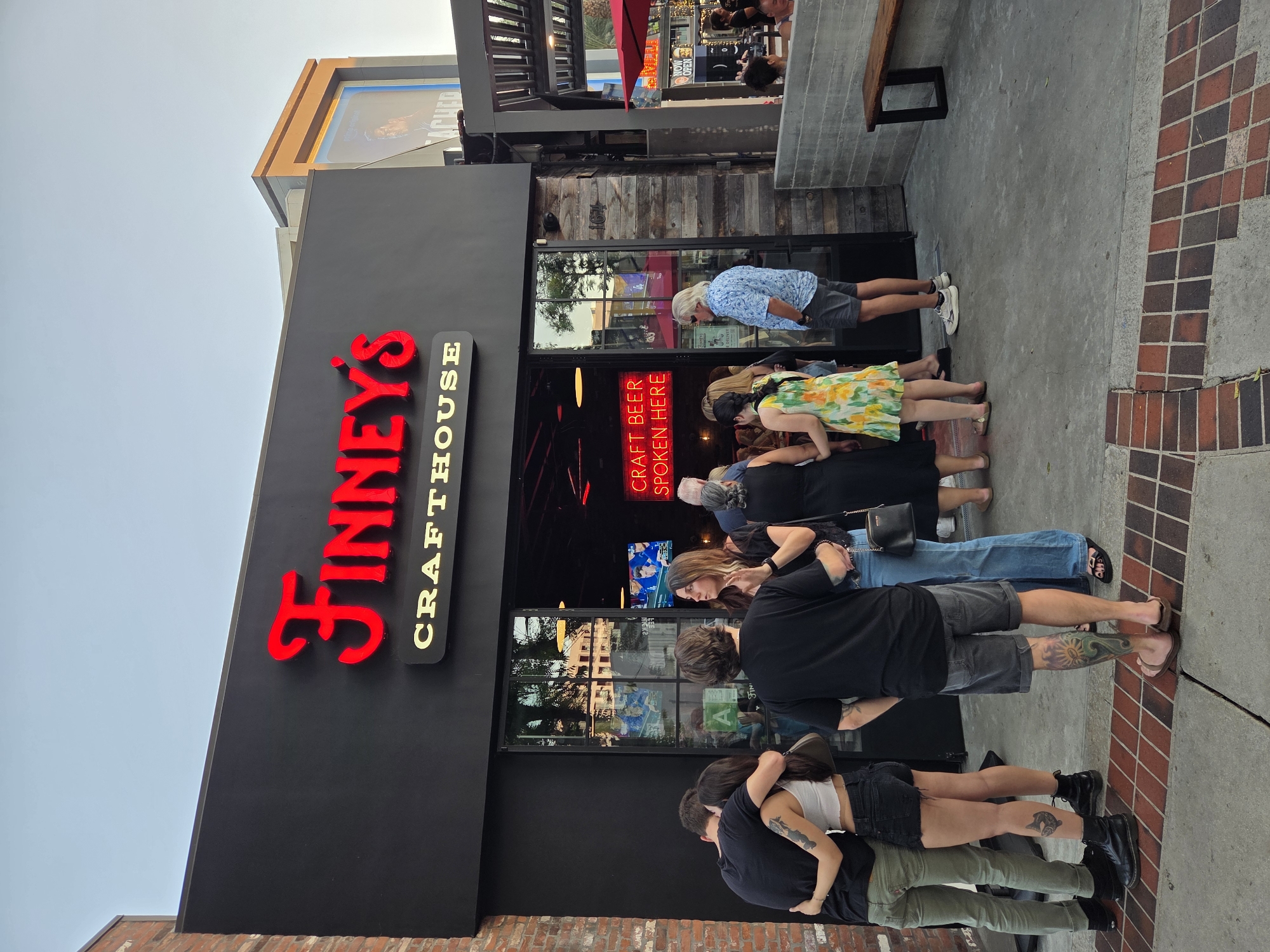 The image depicts the exterior of "Finney's Crafthouse" located in Burbank, California, United States. The building features a black facade with prominent red neon lettering spelling out "Finney's" above "Crafthouse." To the left of the entrance, a group of five young adults is gathered, with two individuals embracing. The individuals are dressed in casual attire, including t-shirts, shorts, and jeans. One person on the far left is wearing green pants, a black t-shirt, and black boots. Next to them, a female is wearing a white crop top, black shorts, and black boots. A third person, also in a black t-shirt, stands with their back to the camera, wearing denim shorts. A woman in a black top and blue jeans is engaged in conversation with another individual. To the right of the entrance, more patrons are visible, including a man in a blue patterned shirt and grey shorts, and a woman in a floral dress. The entrance itself is a large glass door, reflecting the surroundings. Inside the establishment, neon signs are visible, including one that reads "CRAFT BEER SPOKEN HERE." The scene appears to be set during daylight hours with a clear sky. The pavement in front of the building is paved with a mixture of concrete and brickwork. To the right of the building, a shaded outdoor seating area with wooden benches and red umbrellas can be seen, along with additional patrons. In the background, a large digital billboard displays an advertisement.