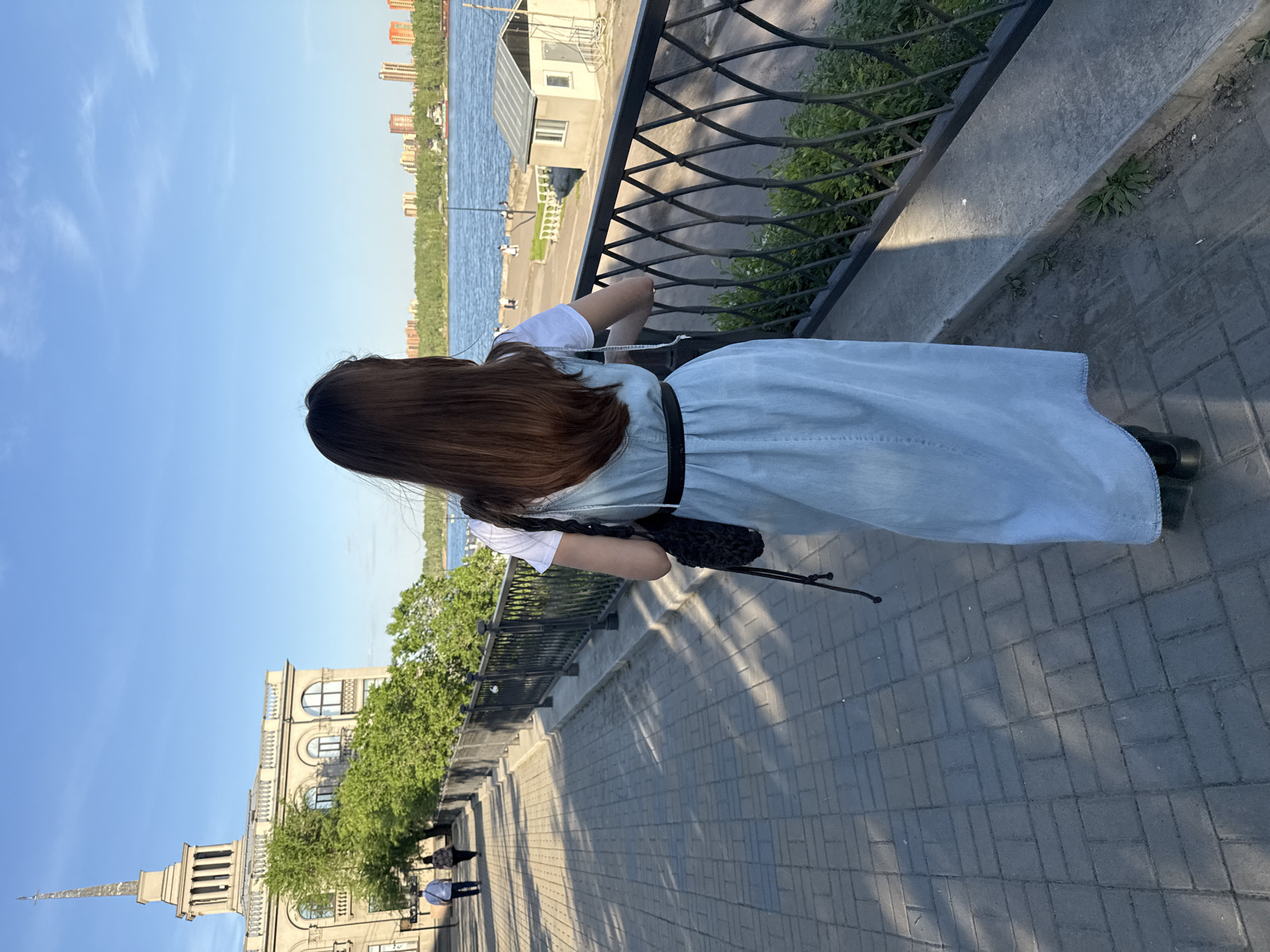 A person is seen from behind, leaning over a metal railing to look out over a wide body of water. The subject has long brown hair and is wearing a white short-sleeved top under a light blue, denim-like pinafore dress with a black belt and black shoes. A black bag hangs from their shoulder.
The scene depicts a sunny day along what appears to be a river embankment or promenade. In the midground, a dark metal railing with a diamond pattern separates a paved walkway from the water. Green foliage is visible on the other side of the railing.
Across the water, the far bank is lined with lush green trees and numerous tall, multi-story buildings, suggesting a cityscape. Closer to the person's left, a large, ornate building with classical architectural elements, including arches, columns, and a prominent spire, stands out. Several other people are visible walking on the paved area near this building. To the person's right, a small, white, utilitarian building with windows is situated close to the water's edge, possibly a dock structure.
The sky is clear blue with a few wispy white clouds. The bright daylight casts distinct shadows, notably from the railing and objects onto the paved walkway, indicating a sunny late afternoon or early morning. There is no visible text in the image. The setting suggests a pleasant day for a stroll along the waterfront, possibly in Krasnoyarsk, Russia, given the architectural style and riverine landscape.