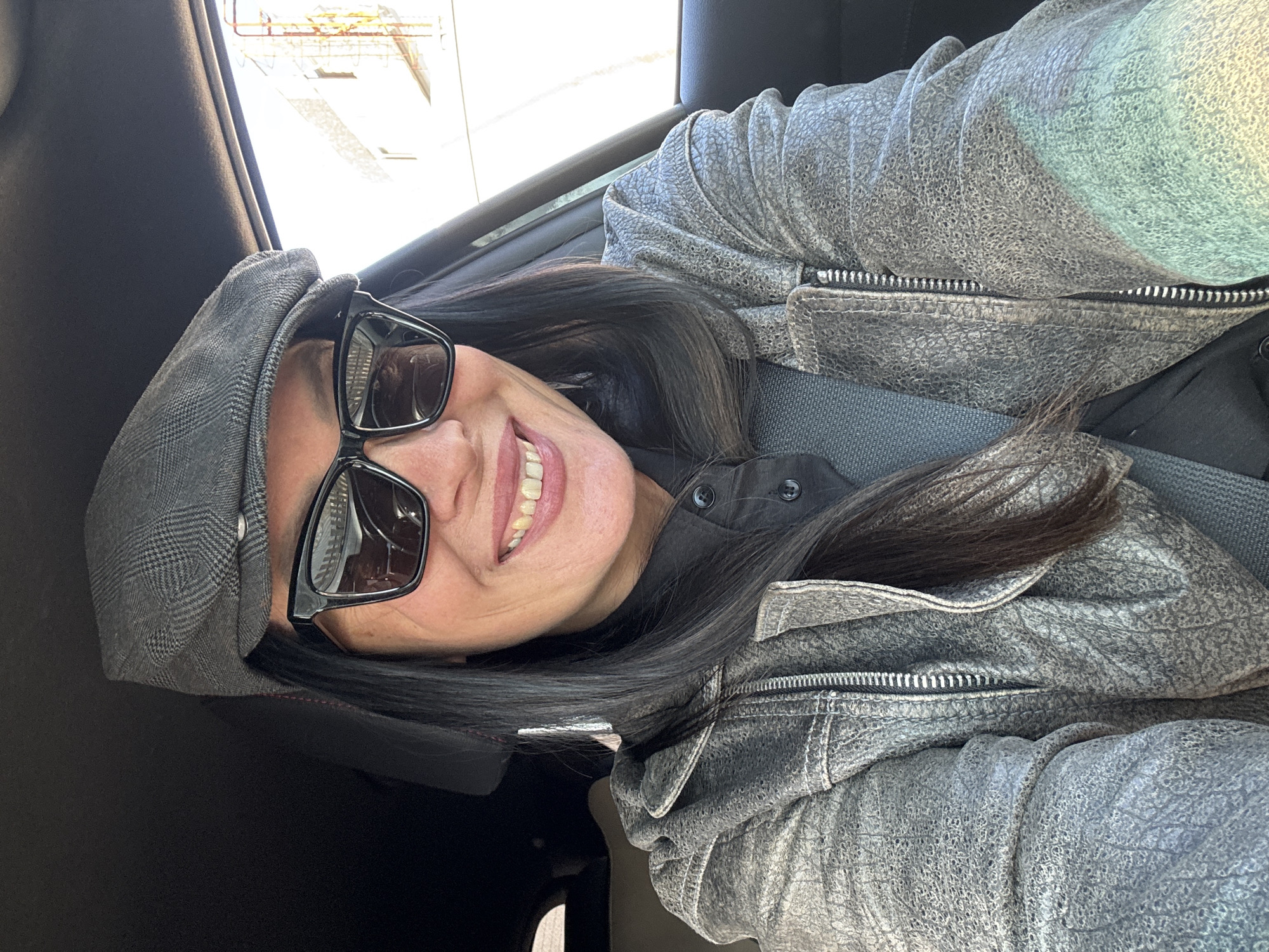 A woman wearing a grey newsboy cap, sunglasses, a black button-down shirt, and a textured grey jacket is seated in a vehicle. She is smiling and secured with a grey seatbelt. The location is indicated as being within Chaikovsky, Russia, but the image does not provide visual confirmation of this location. The interior of the car is visible with a glimpse of the window and surroundings suggesting daylight conditions.