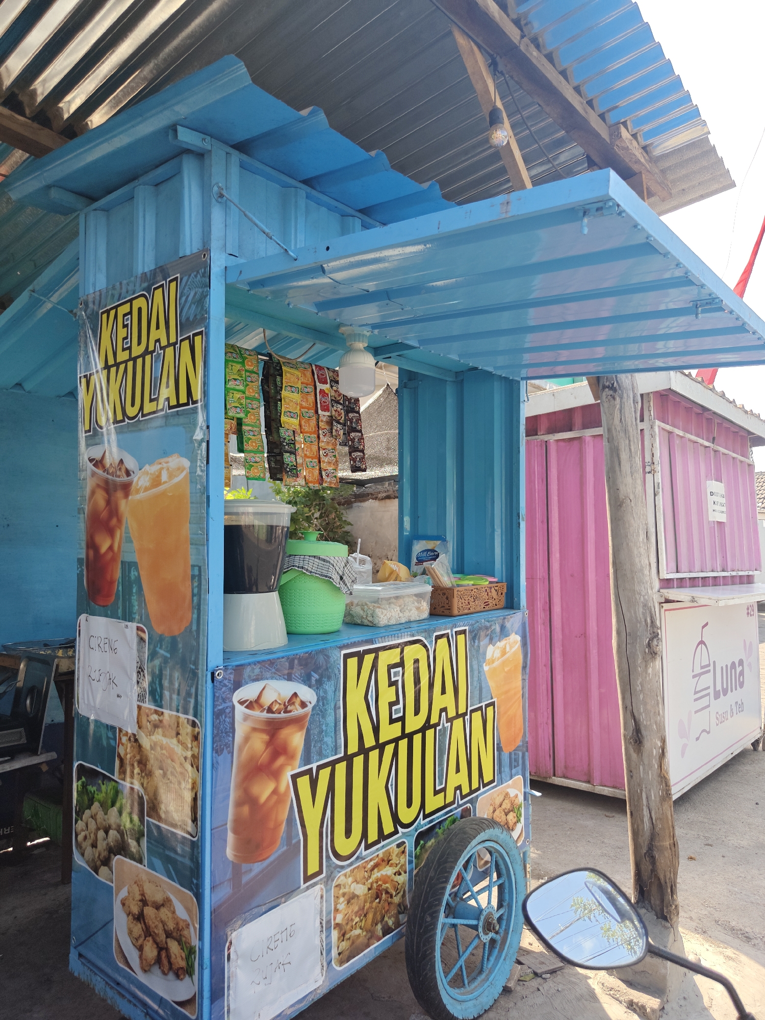 The image depicts an outdoor street food stall, painted predominantly in bright blue. The stall is covered by a blue corrugated metal roof and has a blue corrugated metal body. On the front of the stall, a large banner displays the words "KEDAI YUKULAN" in yellow lettering with black outlines, alongside images of iced beverages and various fried foods. Various packets of snacks are hanging from the front of the stall. To the right, another stall with a pink corrugated metal exterior is partially visible. A wooden pole stands in front of the pink stall. A motorcycle's side mirror, reflecting trees and a sky with clouds, is in the foreground on the right side of the image. The ground appears to be a mix of dirt and broken pavement. The lighting suggests it is daytime, possibly late afternoon, with sunlight casting shadows. The scene is set in an urban or semi-urban environment, typical of street vendors in Indonesia, and is located in the city of Situbondo, Indonesia.