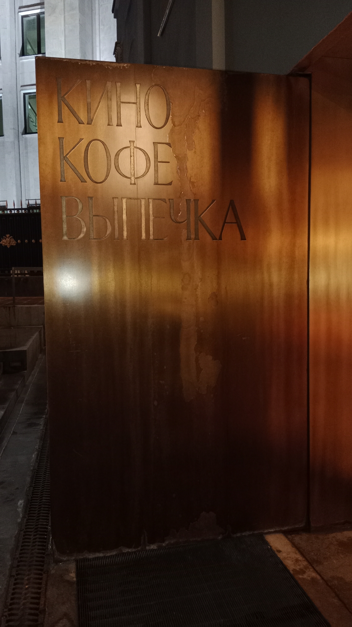 The image shows a sign in Moscow, Russia, with the words "КИНО", "КОФЕ", and "ВЫПЕЧКА" vertically stacked. These translate to "Cinema", "Coffee", and "Pastries" respectively. The sign appears to be made of a bronze or copper-colored metal, and the words are engraved or cut out. The background behind the sign includes a building with windows and a black metal fence. The sign is located at street level. The ground around the sign is a combination of stone and a black metal grate. The lighting is warm, suggesting it is evening or nighttime. There are no people visible in the image.
