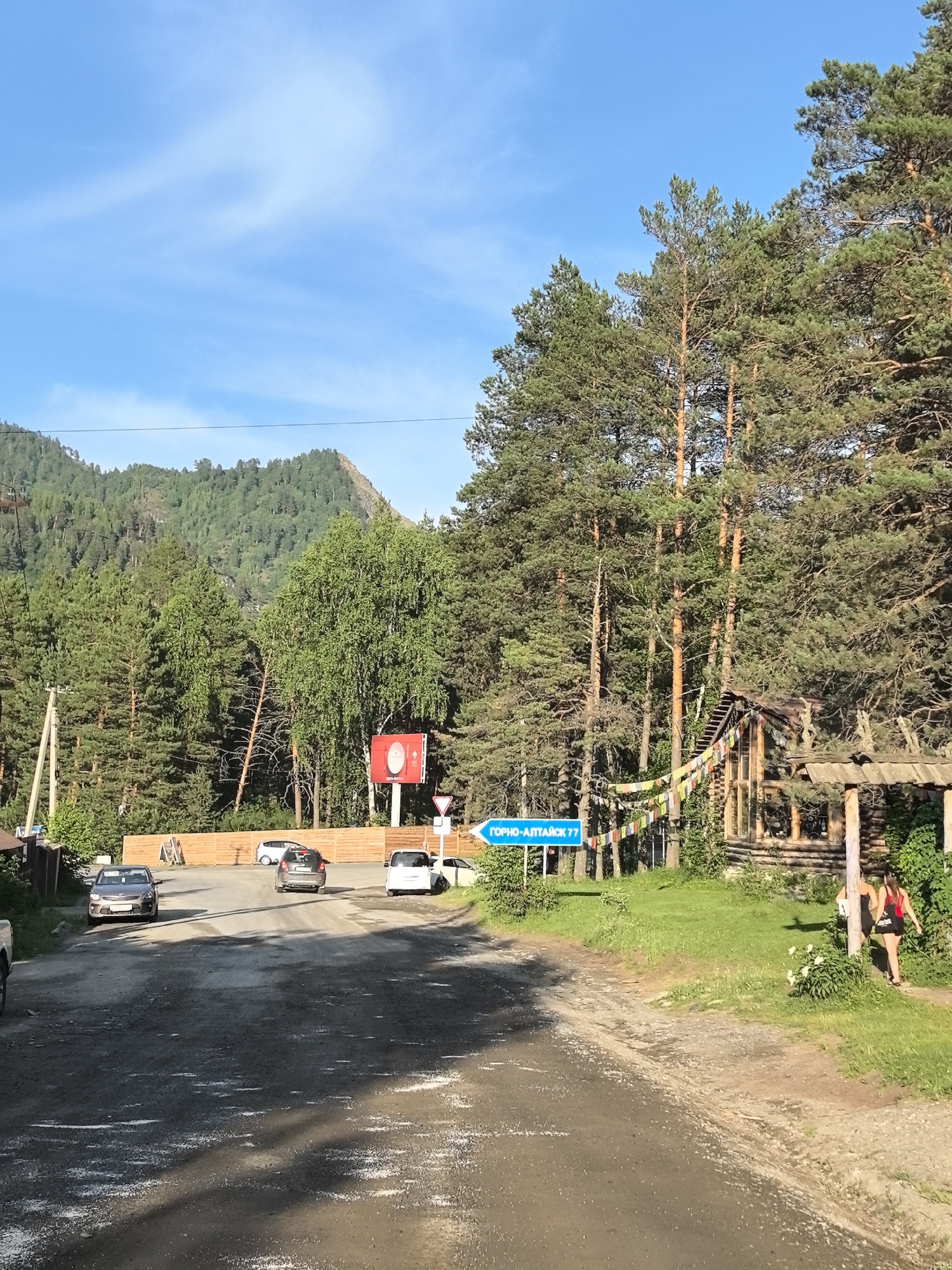 A dirt road curves through a forested area, with a mountain range visible in the background under a clear blue sky with scattered clouds. The road is lined with tall pine trees and deciduous trees. On the right side of the road, there's a rustic wooden structure with prayer flags strung across it, and two people are walking away from the camera. A blue road sign pointing to "GORNO-ALTAISK 77" is also visible. Several cars are parked or driving on the road, and a wooden fence runs along the left side. A red and white billboard is situated behind the fence.
