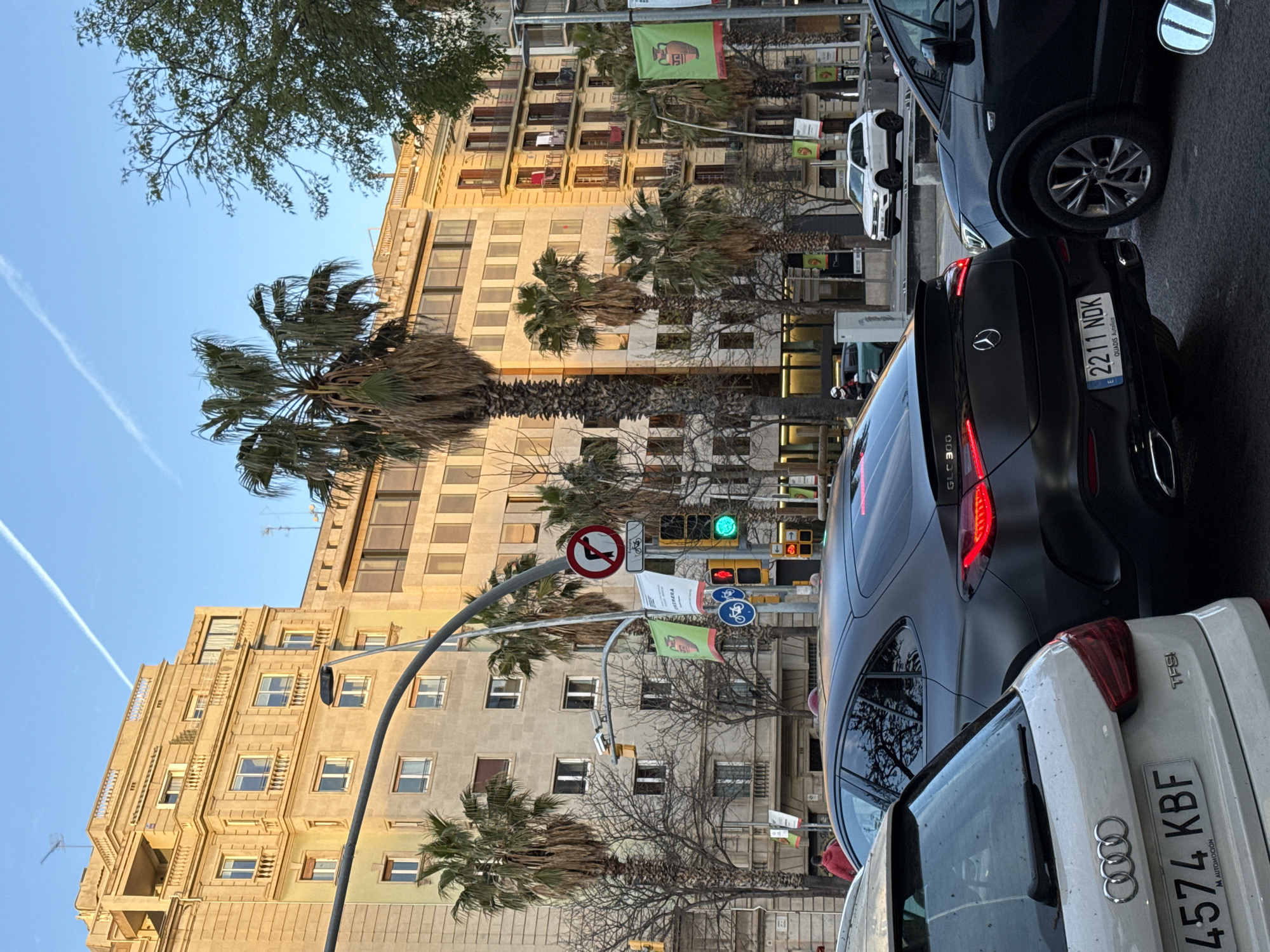 The image depicts a street scene in Barcelona, Spain, under clear blue skies with a prominent white contrail. The time appears to be late afternoon or early evening, given the golden sunlight illuminating the upper facades of the buildings.
In the immediate foreground, the rear of a white Audi A1 hatchback is visible on the left, displaying Spanish license plate "4574 KBF" and "TFSI" badging. Directly ahead of it, occupying the central mid-ground, is the rear of a matte black Mercedes-Benz GLC 300 coupe SUV. Its taillights are illuminated red, and it carries the Spanish license plate "E 2211 NDK". To the right of the Mercedes-Benz, the front-right quarter of a dark-colored SUV is partially visible, showing its multi-spoke wheel. Further down the street on the right, a white SUV, possibly a Volkswagen, is parked or traveling.
The road surface appears wet or reflective. Traffic lights are visible, with the primary vehicular light showing green, indicating active traffic flow. A prominent red circular traffic sign indicating "no left turn" is mounted on a pole, with a supplementary "no entry for bicycles" sign below it. Blue circular signs for bicycle paths are also present. Overhead, a curved grey street lamp arc spans the intersection, with security cameras mounted on a pole.
The background consists of multi-story, traditional Barcelona-style apartment buildings. The building on the left features a light-colored, ornate facade with numerous windows and balconies, basking in the sunlight. Numerous palm trees, some tall and mature, line the street, interspersed with bare deciduous trees. A person in a red hoodie is visible walking on the sidewalk behind the Mercedes-Benz on the left. On the right side of the street, another building with darker, more weathered balconies has laundry hanging from one of its upper levels. Green banners, one featuring an image of an ancient amphora, hang from lampposts along the street.