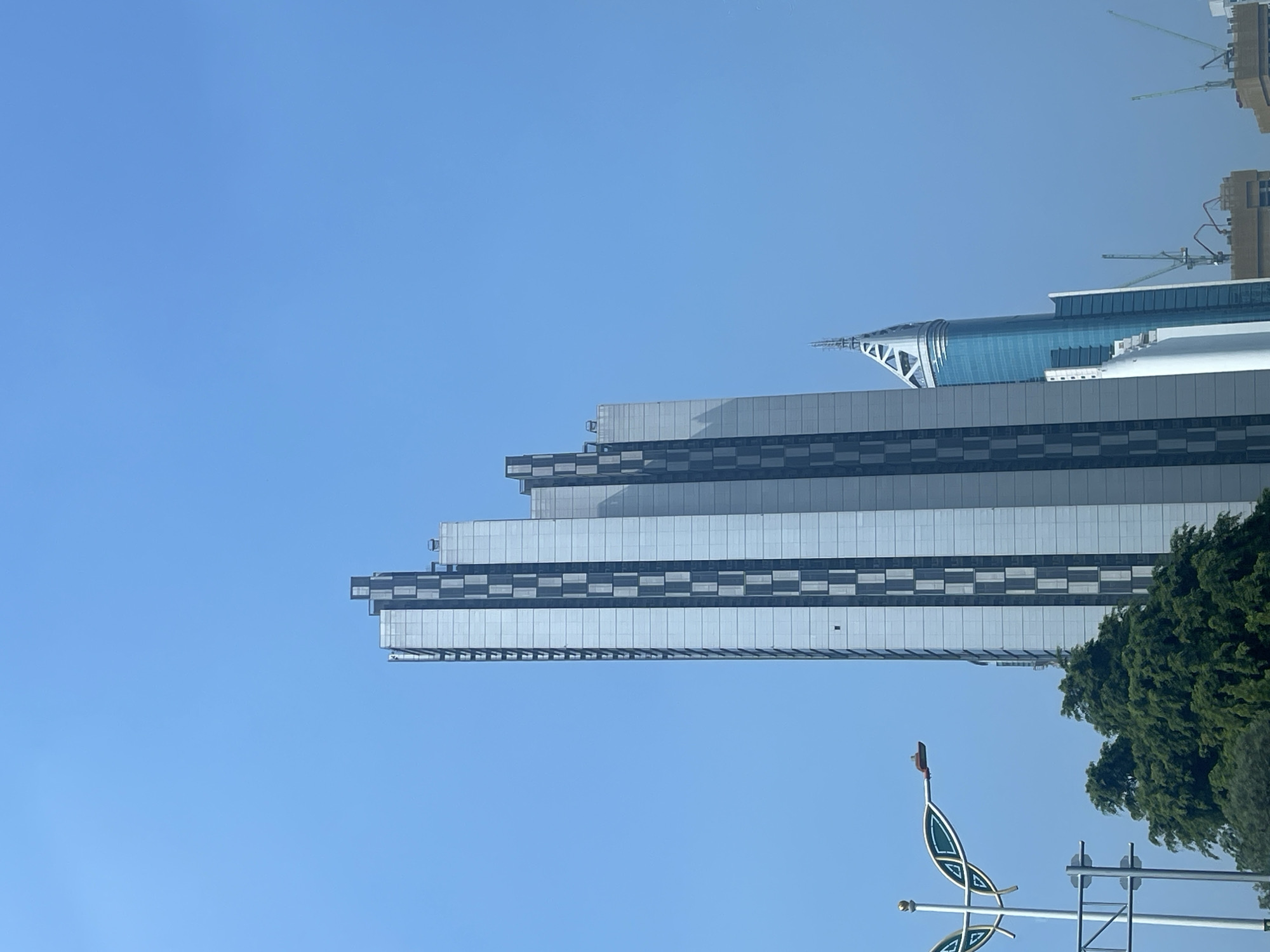 The image depicts a clear blue sky over a city skyline. In the foreground, a tall, modern skyscraper with a silver facade and dark vertical accents dominates the center-left of the frame. To the right of this building, a second, narrower skyscraper with a blue glass exterior and a pointed, white crown is visible. In the lower left quadrant, the top of a lush green tree is present, partially obscuring a decorative street lamp and traffic signals. The overall impression is one of urban development set against a bright, cloudless day. This scene is consistent with the modern architecture found in Central Jakarta, Indonesia, a district known for its high-rise buildings and commercial centers.