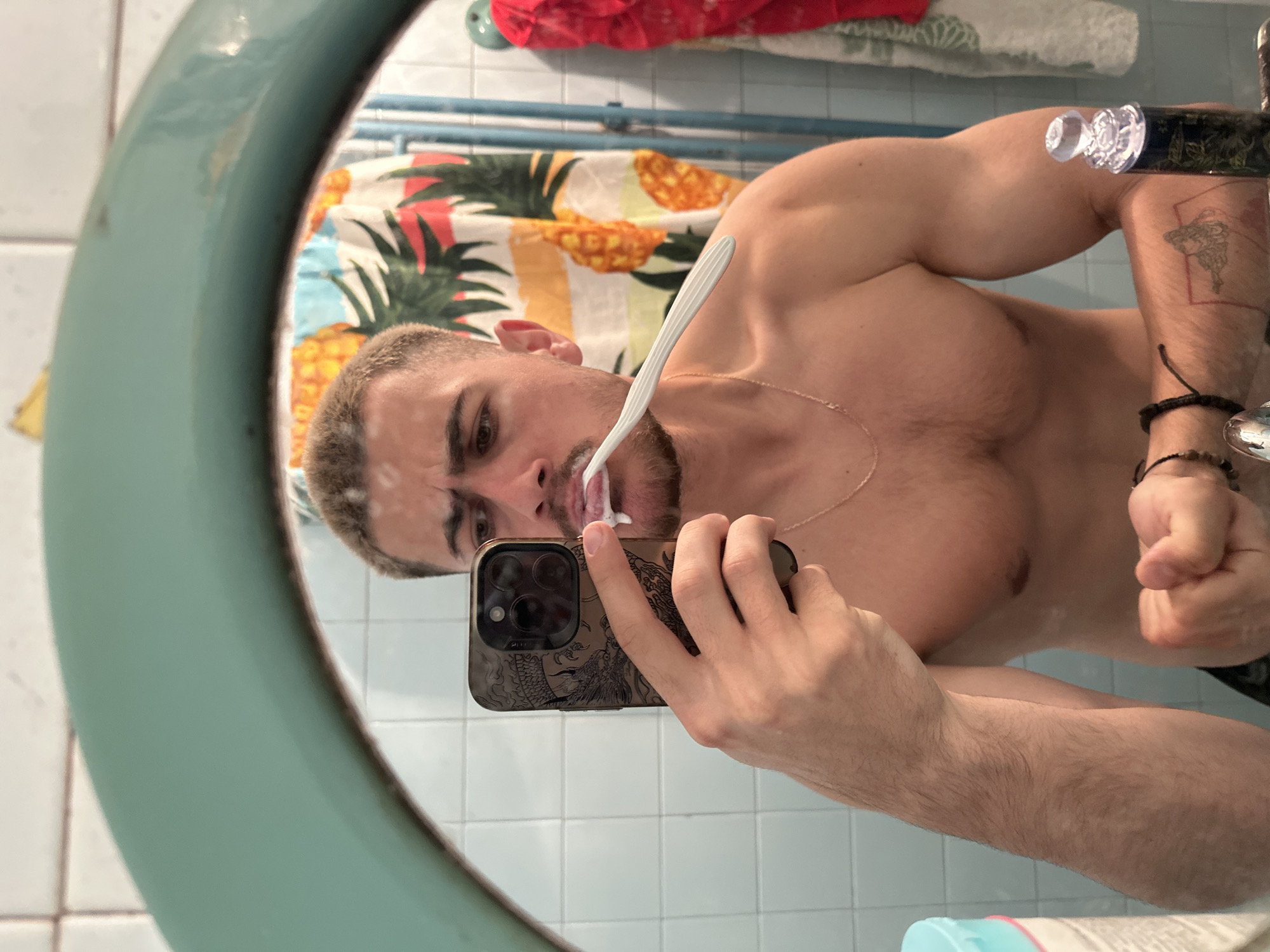 A shirtless man is brushing his teeth while taking a selfie in a bathroom mirror. The mirror has a light blue frame. He is fair-skinned with short, light hair and a short beard. He is wearing a thin gold chain around his neck. He is holding an iPhone with a decorative case. A pineapple-themed shower curtain is visible in the background. The bathroom has white tiled walls and a blue towel rack. Burgas, Bulgaria is not ascertainable from the image.