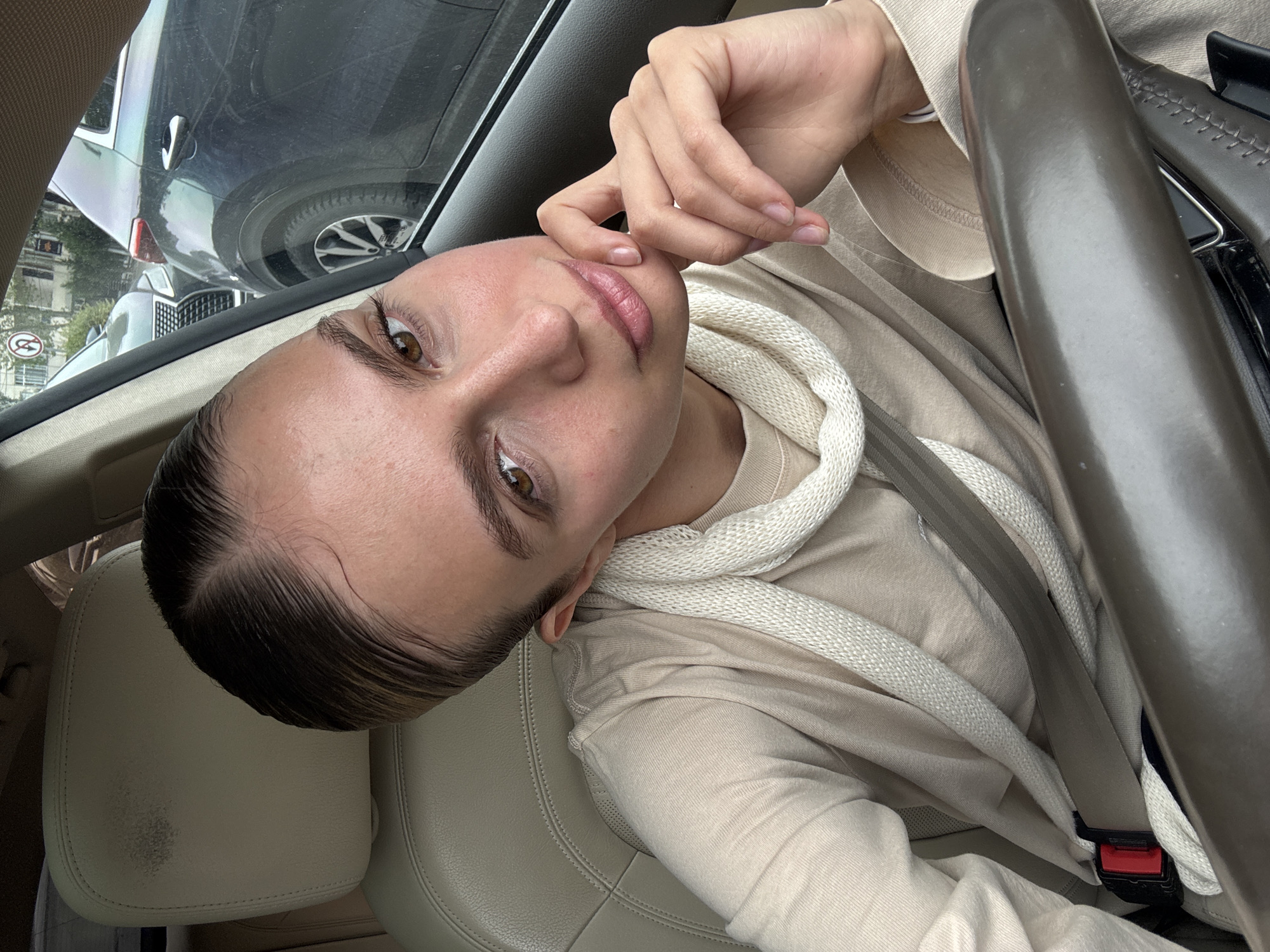 A person with light skin, brown eyes, and light brown hair is sitting in the driver's seat of a car. They are wearing a light beige hooded sweatshirt and a seatbelt. The person's left hand is near their mouth. The steering wheel is on the right side of the frame. Outside the car window, other vehicles are visible, parked on a street in a city. There is a street sign visible, indicating the image was taken in Unknown, Unknown.