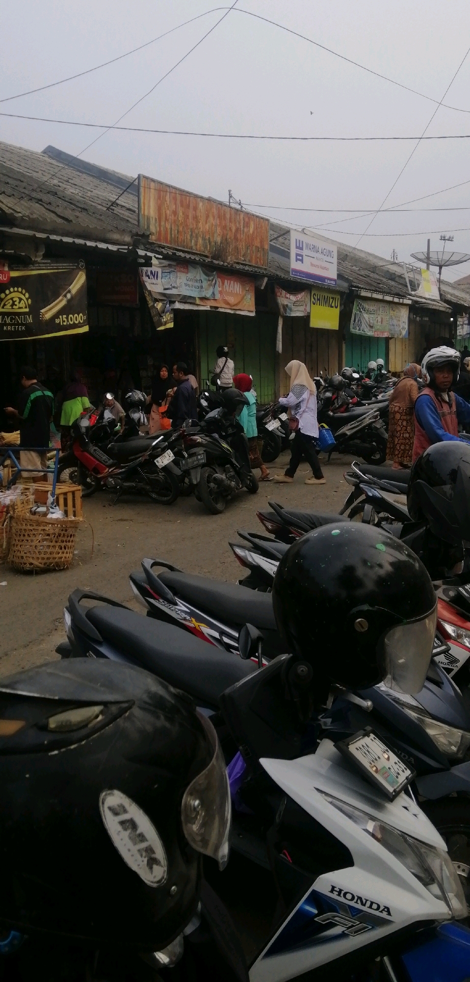 The image depicts a street scene in Ajibarang, Indonesia, likely a market or commercial area, characterized by a row of shops or stalls. The foreground and middle ground are dominated by a large number of parked motorcycles, with several more visible in the background. The motorcycles are predominantly scooters and smaller displacement bikes, suggesting a common mode of transportation.
Several people are visible in the image, most of whom appear to be customers or vendors. In the mid-ground, a woman in a hijab is walking past a line of motorcycles, carrying a blue bag. To her left, another woman in a hijab and a patterned skirt is walking away from the camera. A man in a blue shirt and a red head covering is walking towards the right, and a man in a dark shirt and light pants is standing near a stall. Further to the left, a person in a green shirt is interacting with someone behind a stall, and another person in a dark green jacket is also present. In the far right of the middle ground, a person wearing a helmet and a blue jacket is visible.
The shops or stalls have corrugated metal roofs and some have awnings or banners displaying advertisements. Notable signs visible include "MAGNUM KRETEK Rp 15.000," "WARNA AGUNG PAINTS," and "SHIMIZU." The ground is paved or made of packed earth. The sky is overcast, contributing to a subdued lighting condition.
In the foreground, two motorcycle helmets are prominently displayed. One is a black helmet with a clear visor and the brand "INK" visible on the side. It is placed on a scooter with white and blue bodywork, featuring the "HONDA F1" logo. Another black helmet is visible to the left, partially obscuring the view of the motorcycle it is on. A wicker basket filled with goods is situated to the left of the parked motorcycles. The overall impression is one of a busy, everyday urban environment in Ajibarang.
